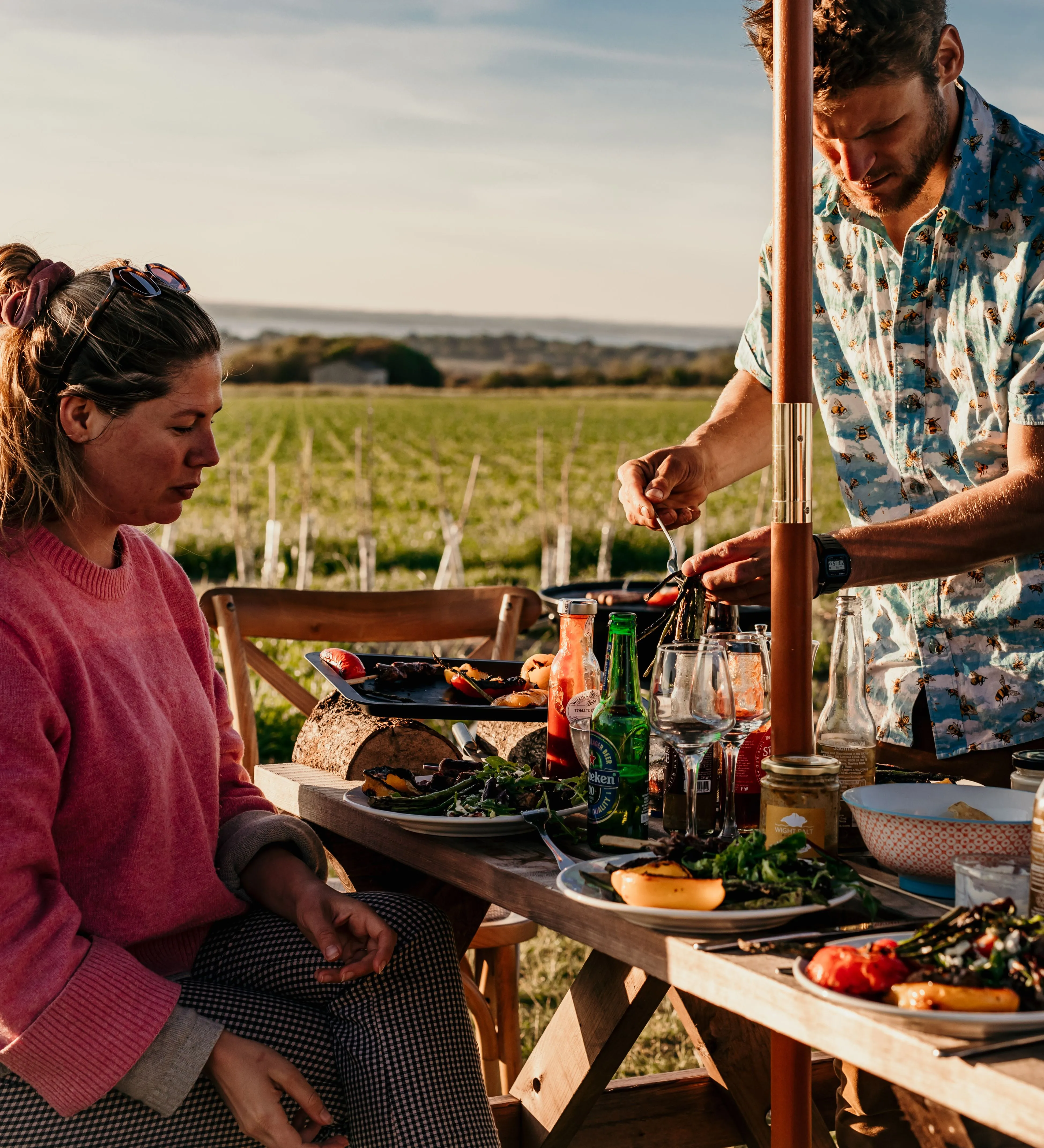 Two people enjoying an outdoor barbecue meal at a wooden picnic table in a countryside field at sunset.