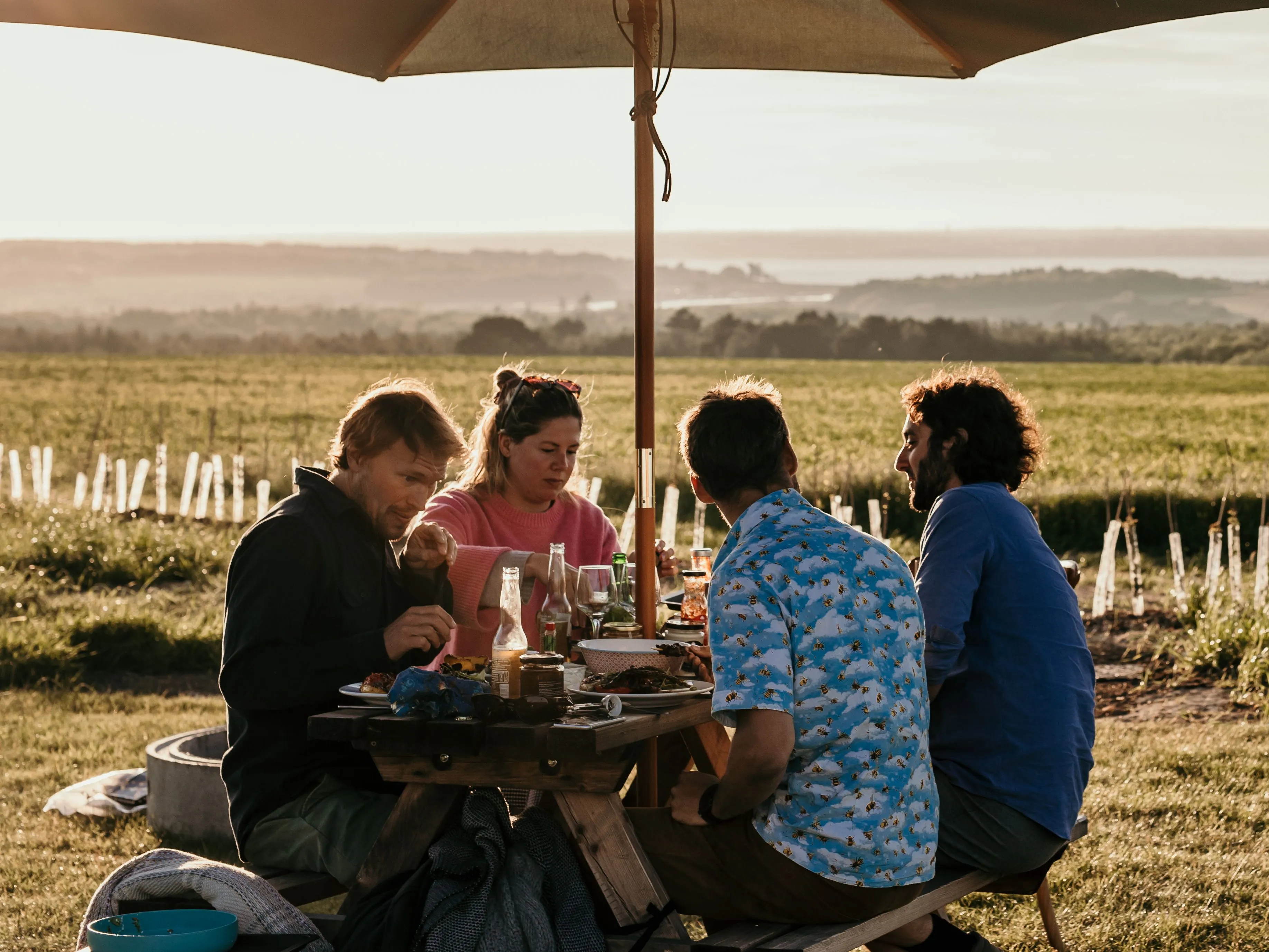 Friends eating outdoors at a picnic table under a large parasol beside a barbecue in a countryside field at sunset.