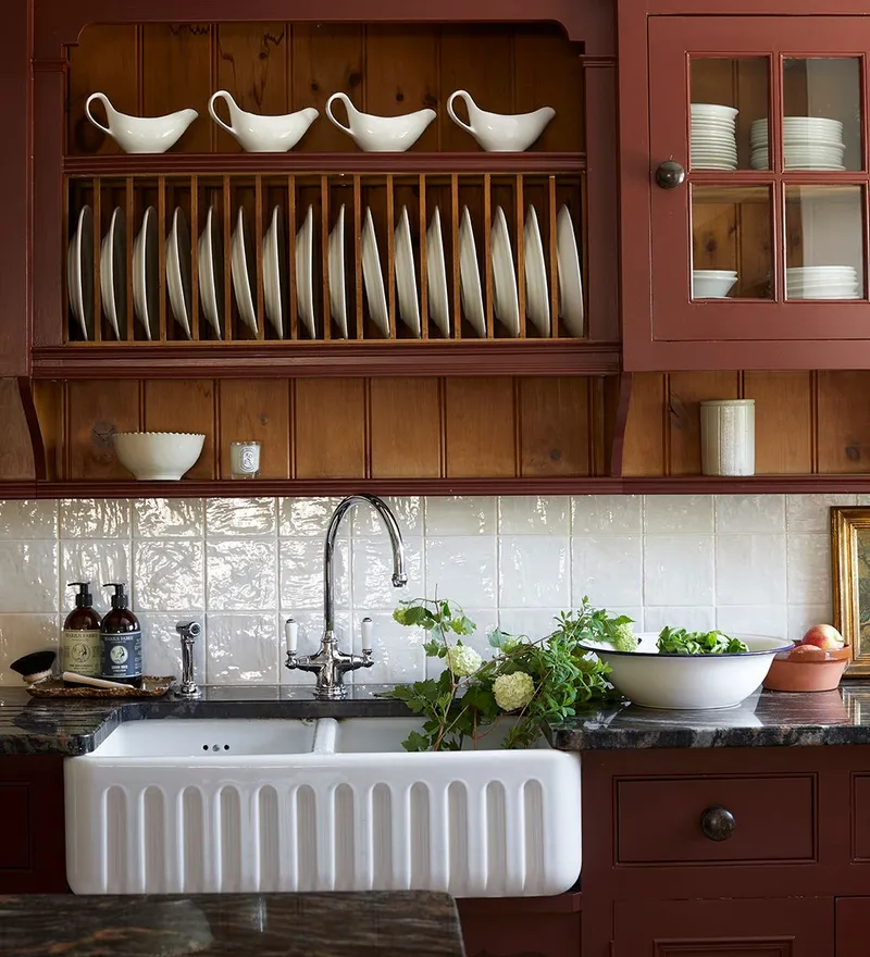 Farmhouse kitchen sink with burgundy cabinetry, open plate rack and white crockery on wooden shelves.