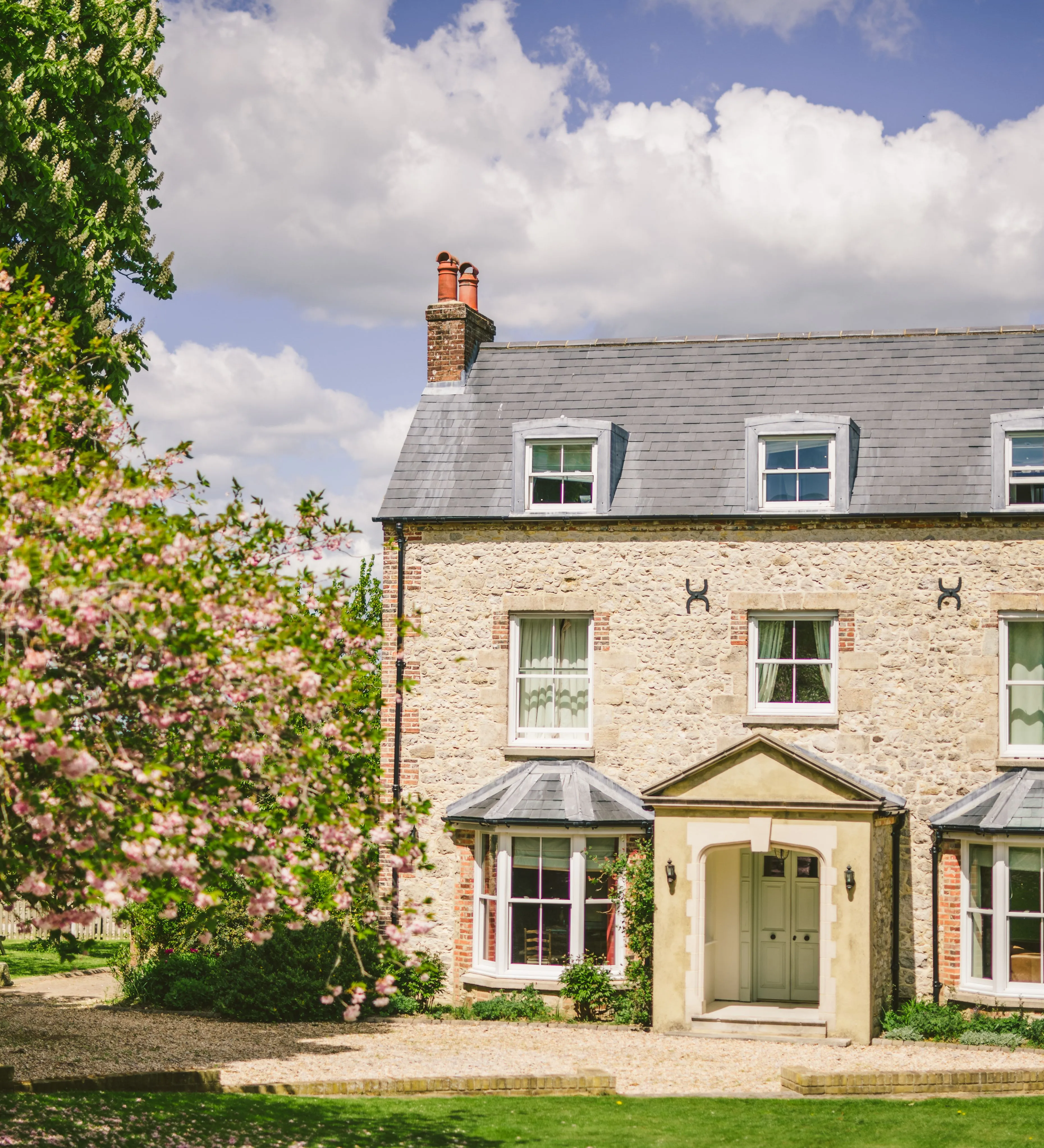 Stone farmhouse with slate roof and dormer windows, framed by a blossoming tree under a blue sky