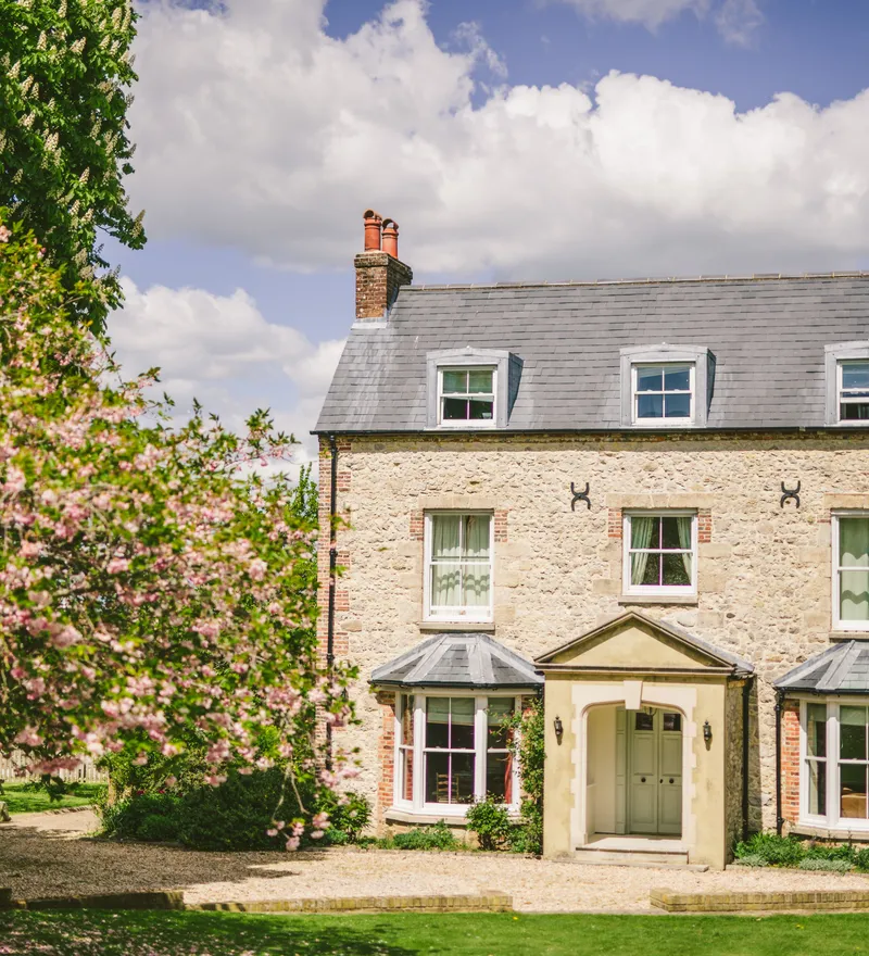 Stone farmhouse with slate roof and dormer windows, framed by a blossoming tree under a blue sky