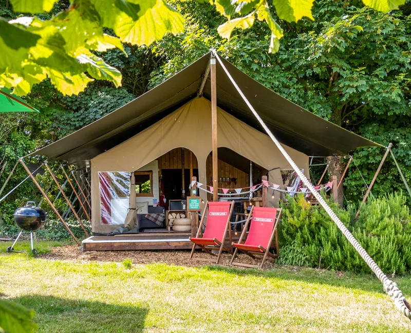 Safari-style glamping tent with a covered porch, surrounded by greenery, deck chairs, and a barbecue grill.