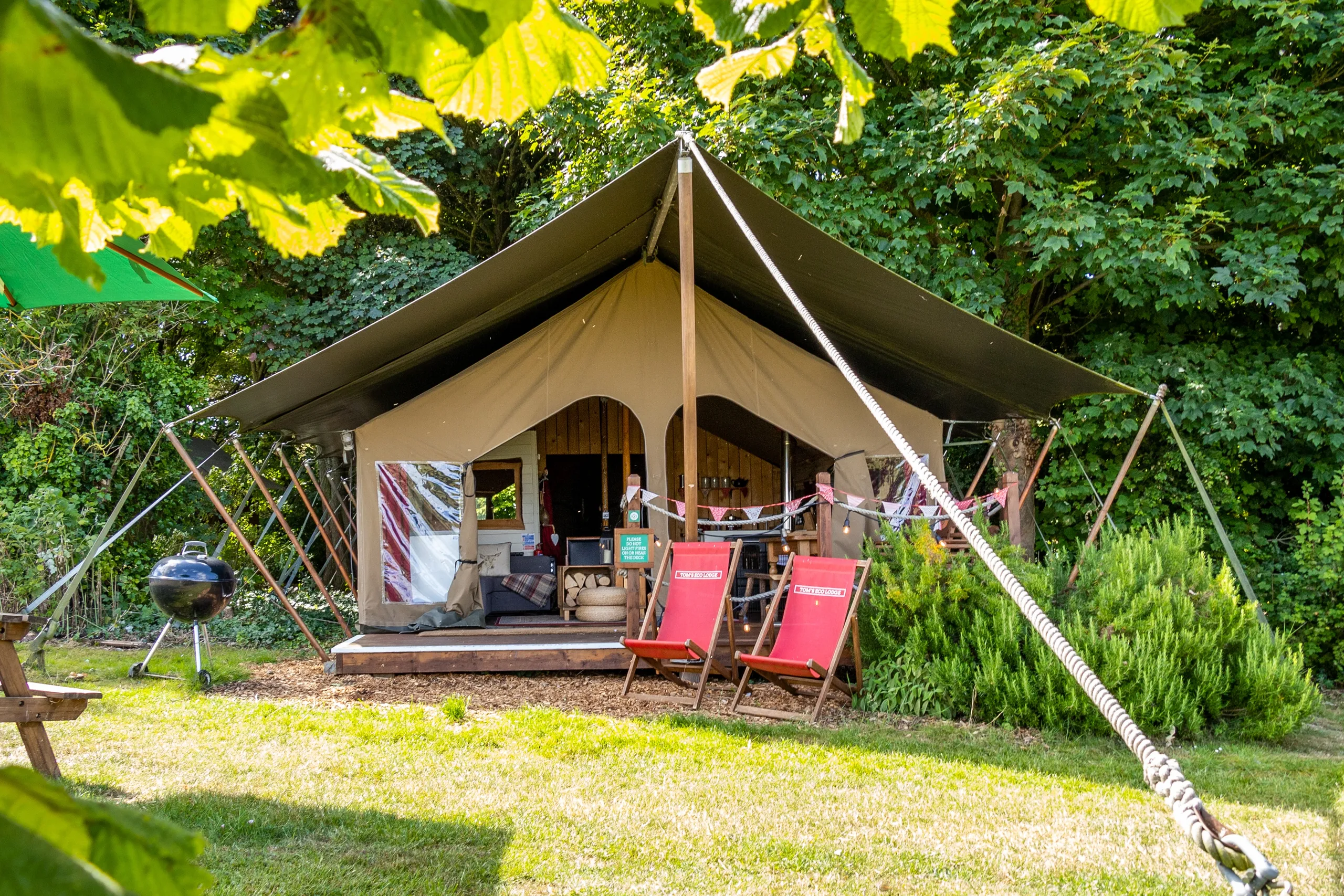 Safari-style glamping tent with a covered porch, surrounded by greenery, deck chairs, and a barbecue grill.