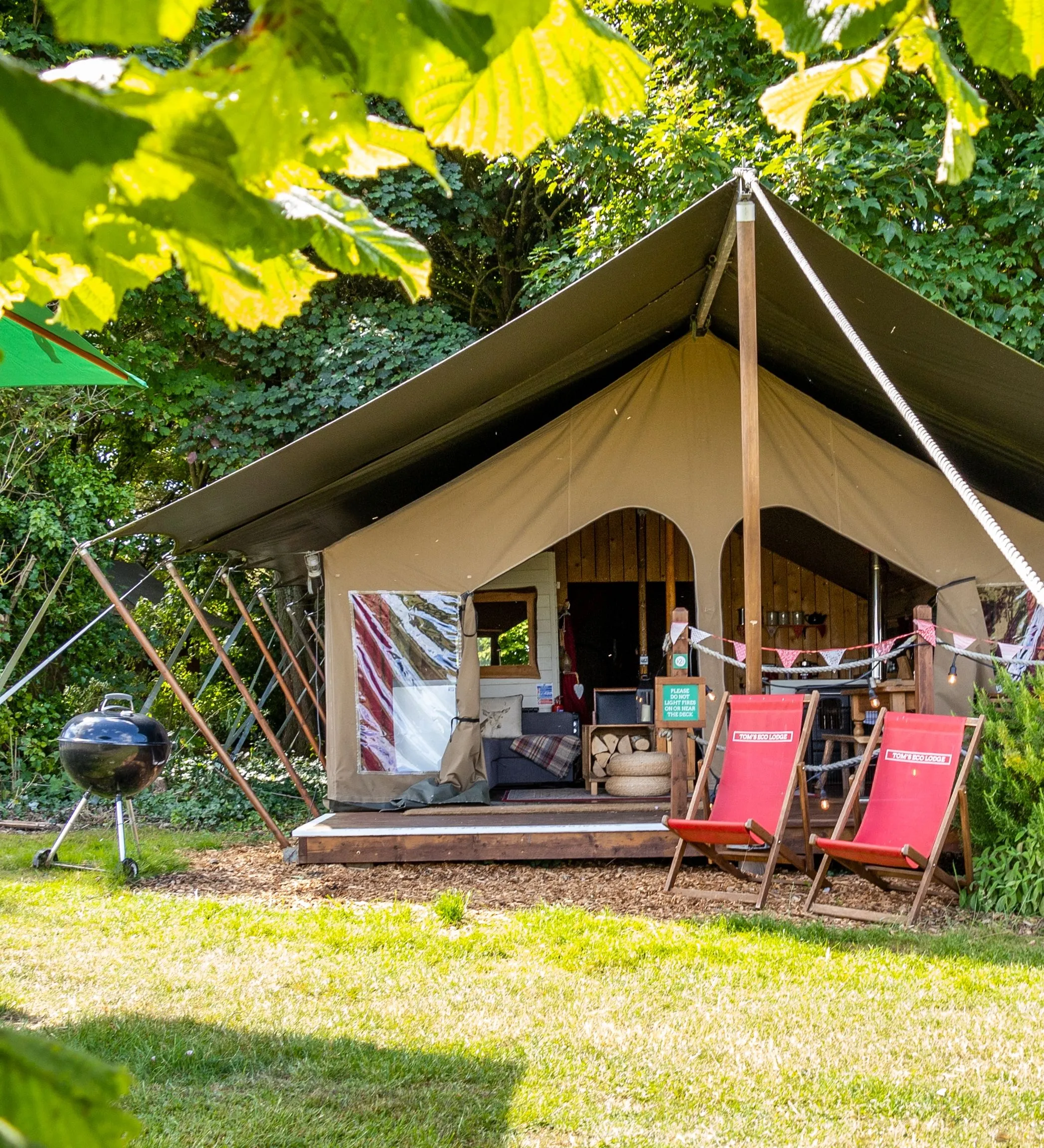Safari-style glamping tent with a covered porch, surrounded by greenery, deck chairs, and a barbecue grill.