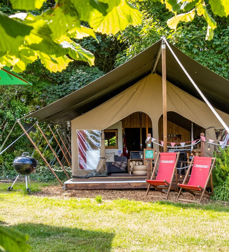 Safari-style glamping tent with a covered porch, surrounded by greenery, deck chairs, and a barbecue grill.