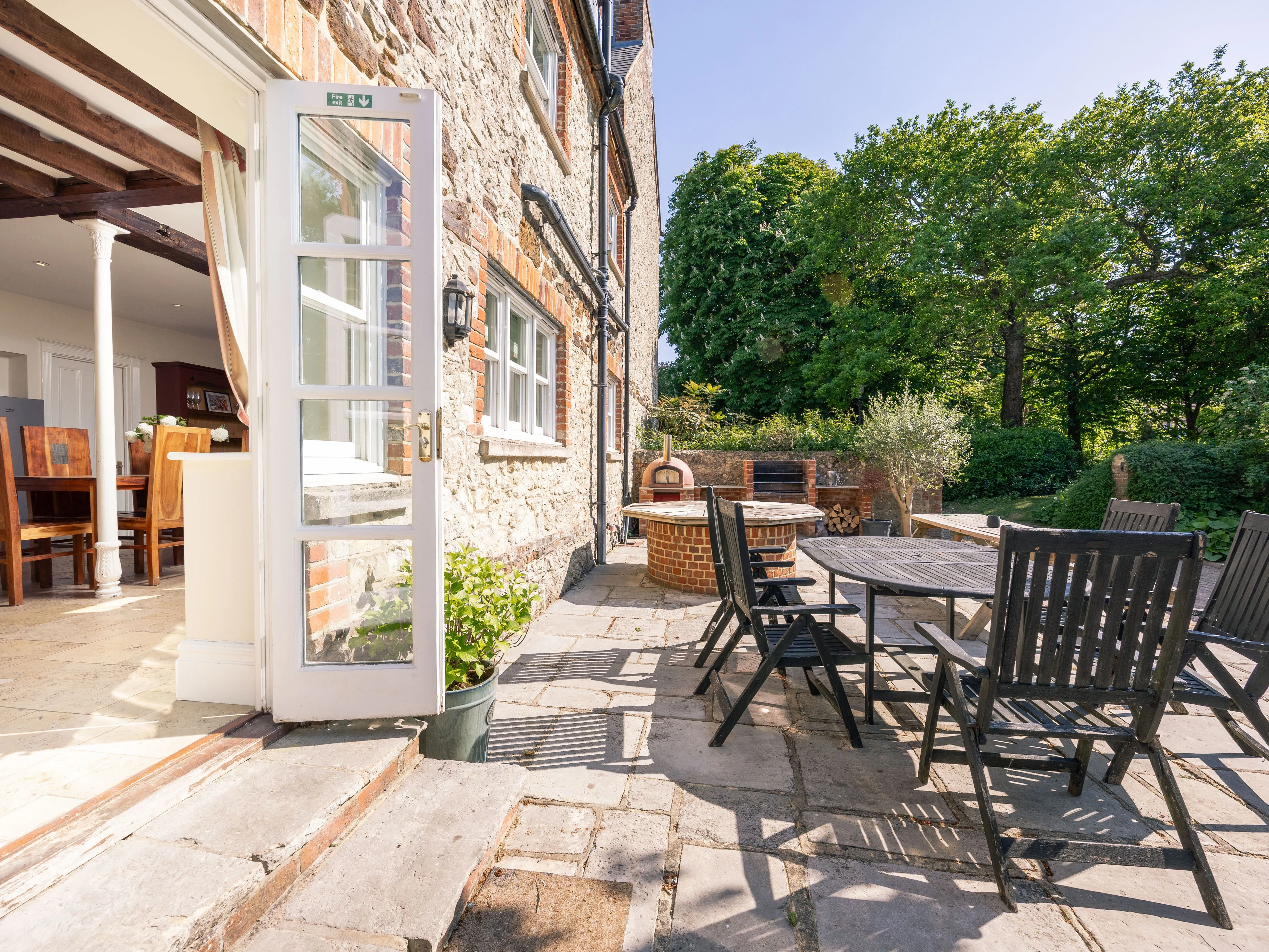Open French door from farmhouse kitchen to sunlit stone patio with outdoor dining table and pizza oven