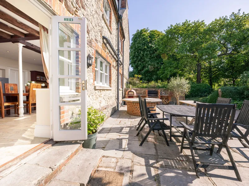 Open French door from farmhouse kitchen to sunlit stone patio with outdoor dining table and pizza oven