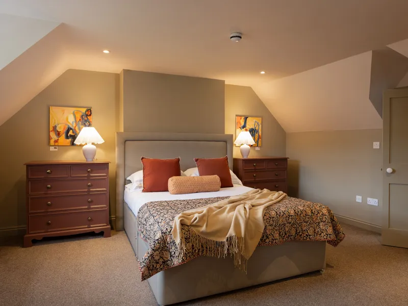 Cosy attic bedroom with upholstered double bed, patterned throw, bedside lamps and chest of drawers