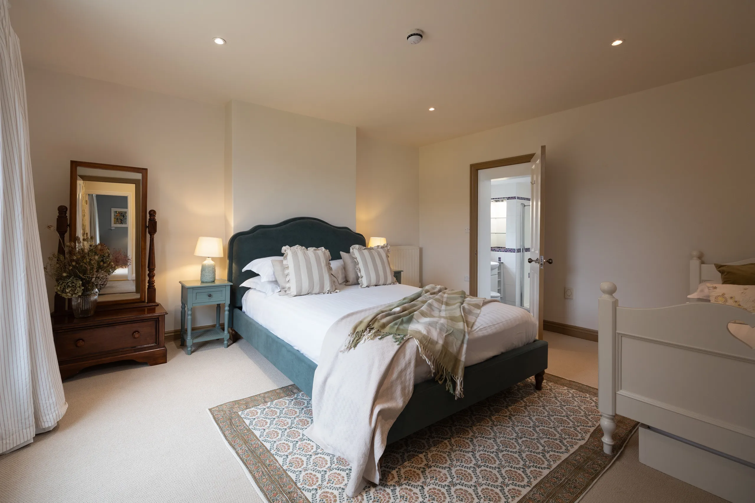 Bright farmhouse bedroom with teal upholstered double bed, bedside lamps, wooden dressing table and doorway to en-suite bathroom