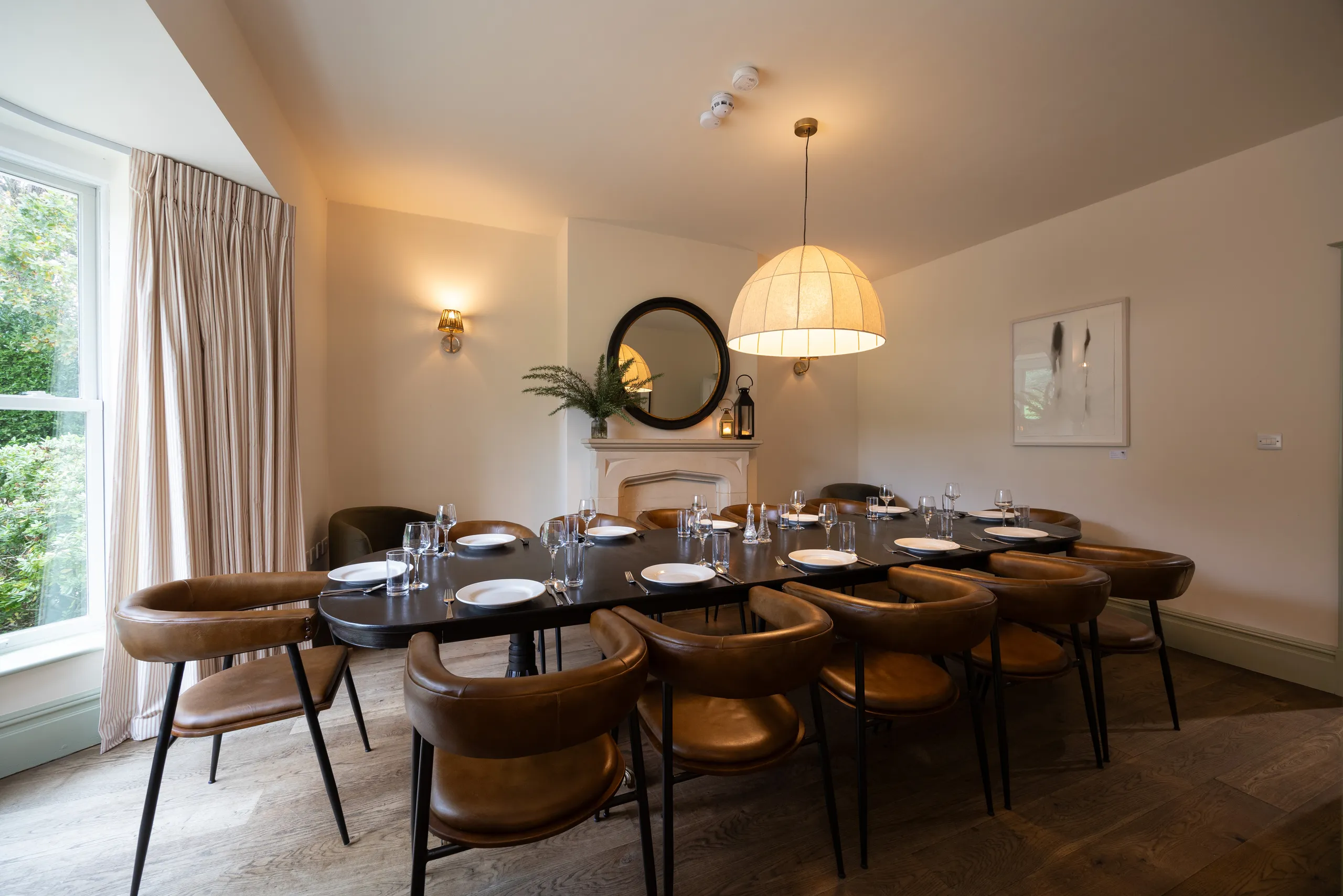 Dining room with a long table set for a meal, leather chairs, and a large pendant light beside a window with curtains