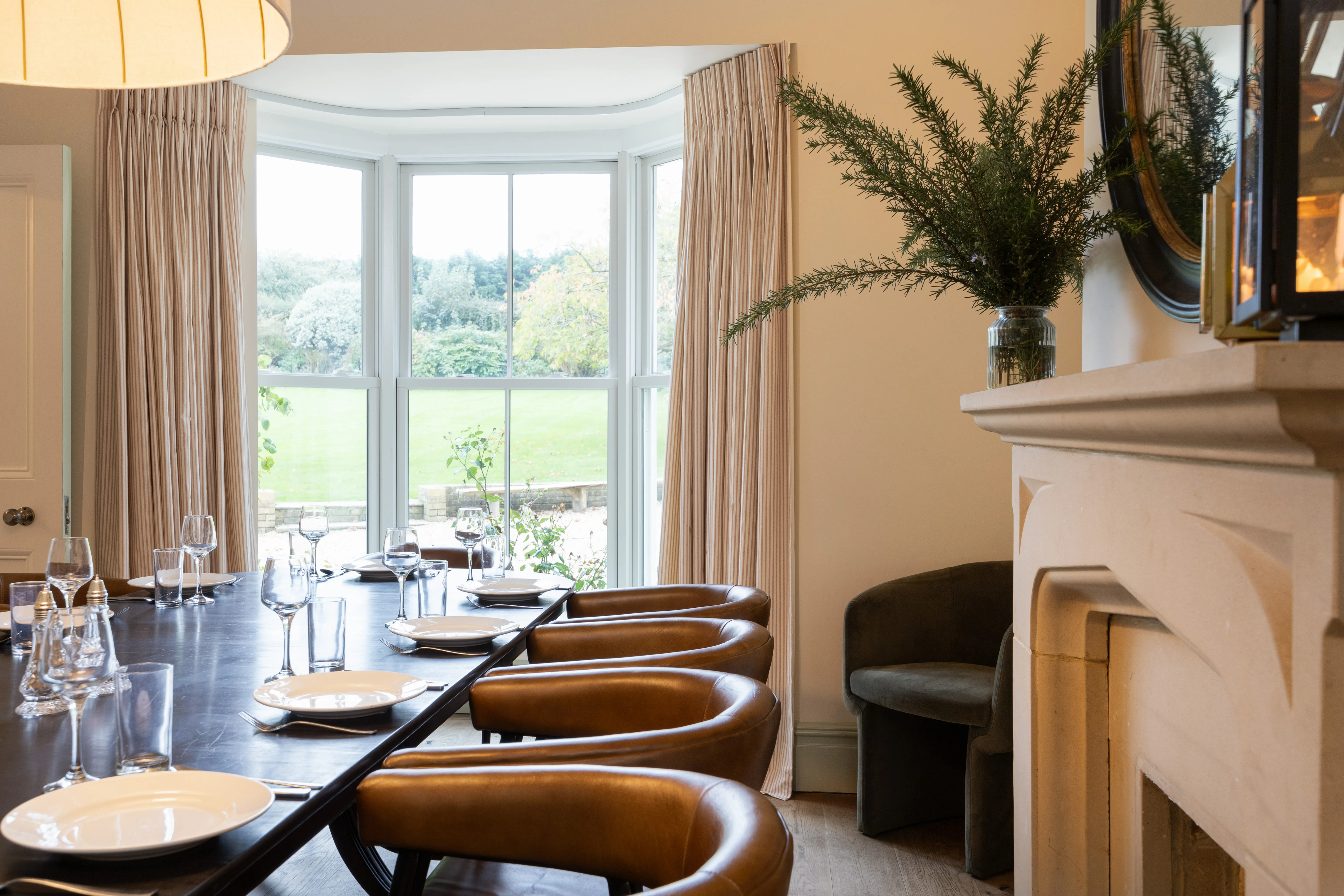 Dining room table set for a meal beside a large bay window with curtains and leather chairs, overlooking a garden.