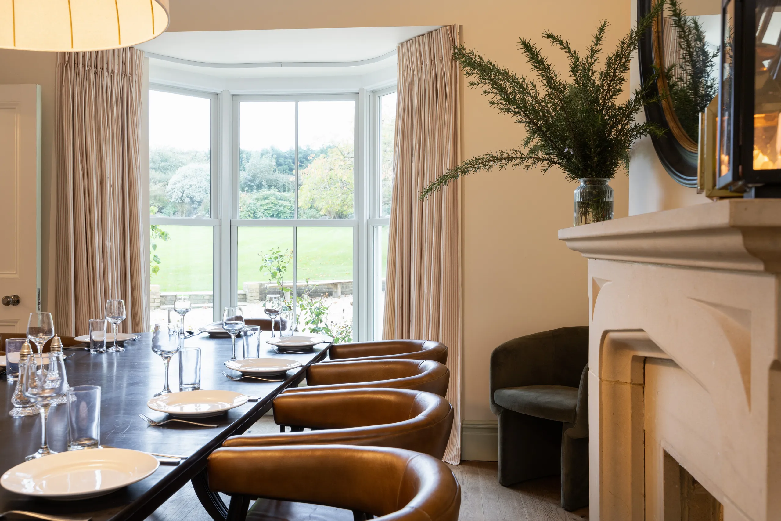 Dining room table set for a meal beside a large bay window with curtains and leather chairs, overlooking a garden.