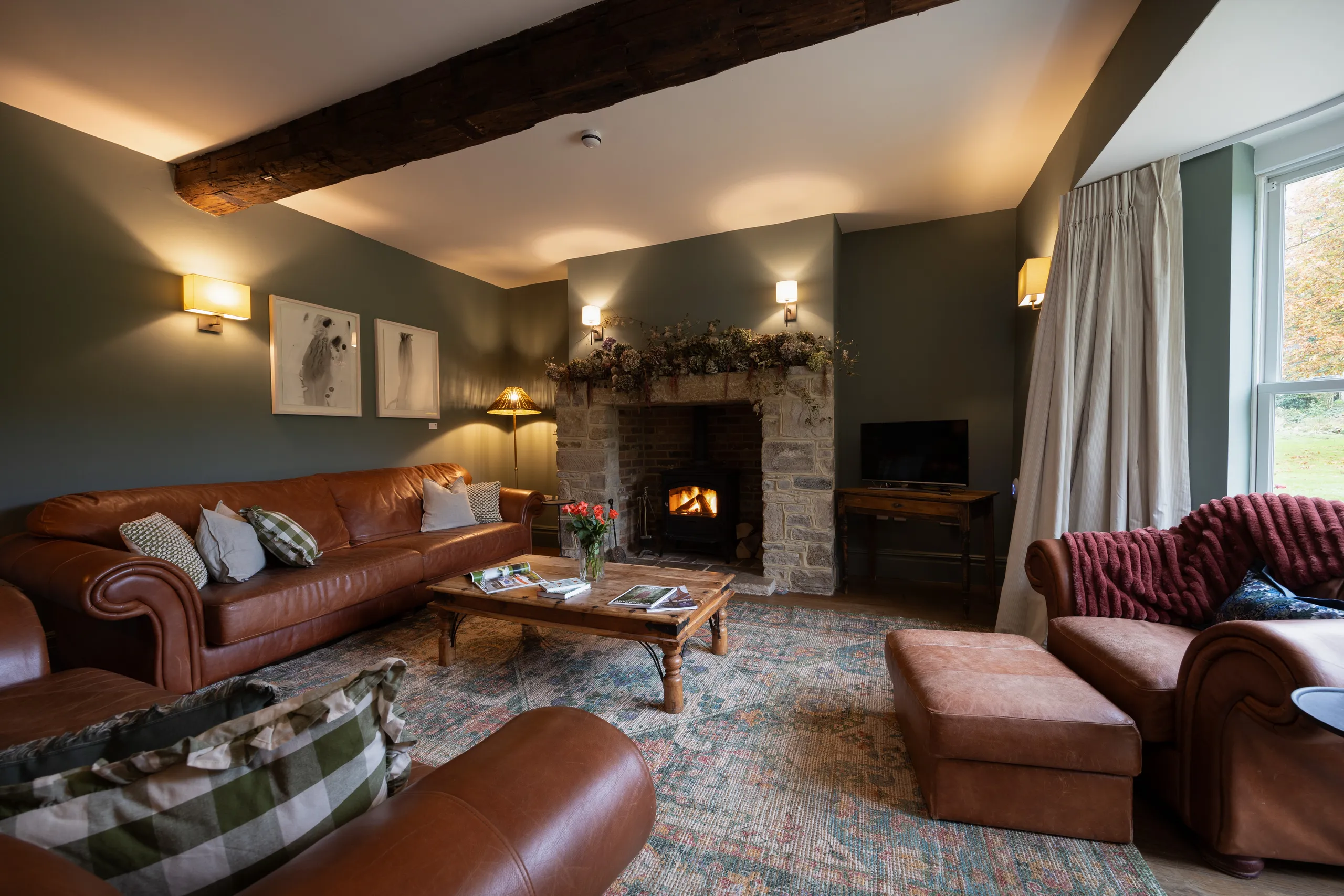 Cosy farmhouse sitting room with leather sofas, wood-burning stove in a stone fireplace, and a large window with curtains.