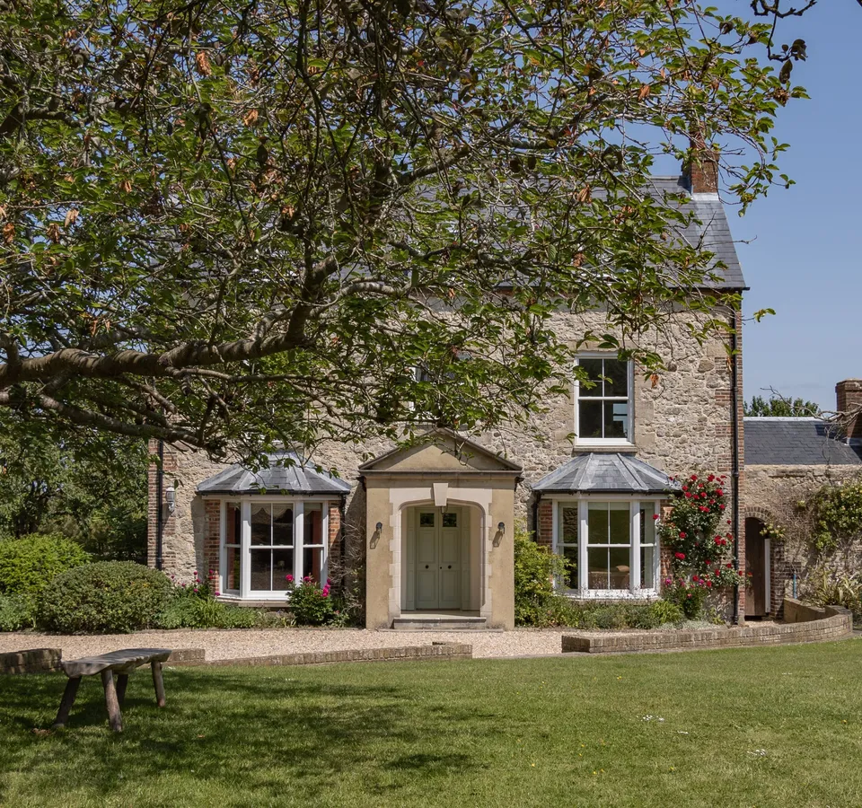Stone farmhouse with bay windows and a central porch, viewed across a lawn beneath a large tree