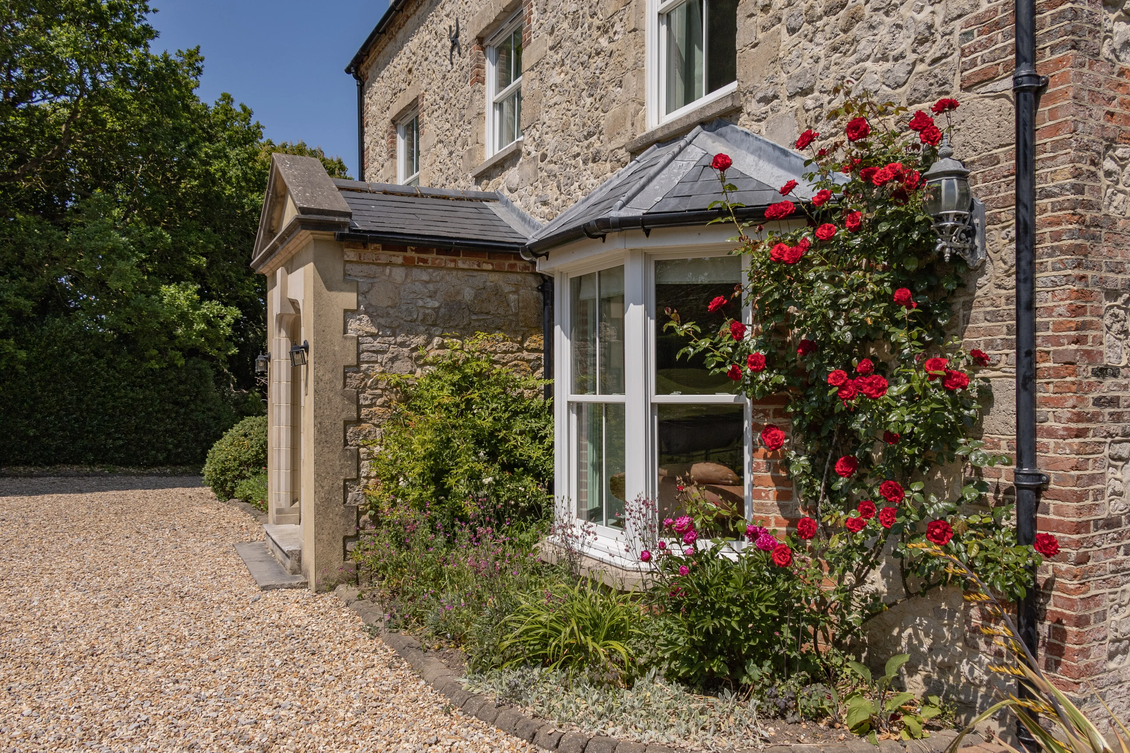 Stone farmhouse exterior with bay window and climbing red roses beside a gravel driveway