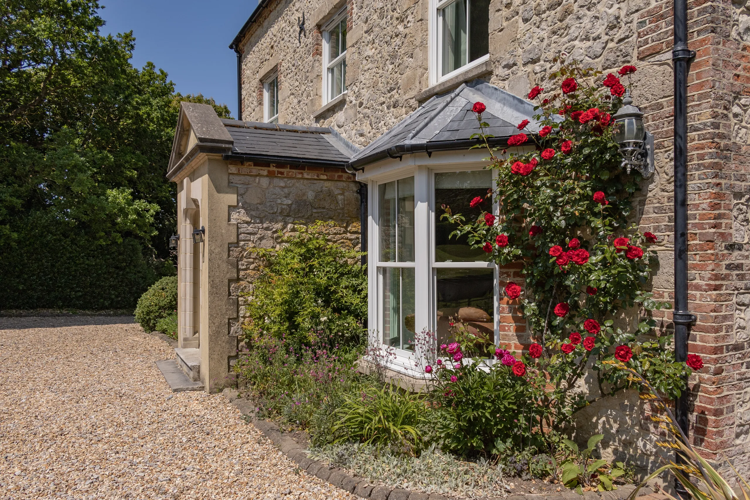 Stone farmhouse exterior with bay window and climbing red roses beside a gravel driveway