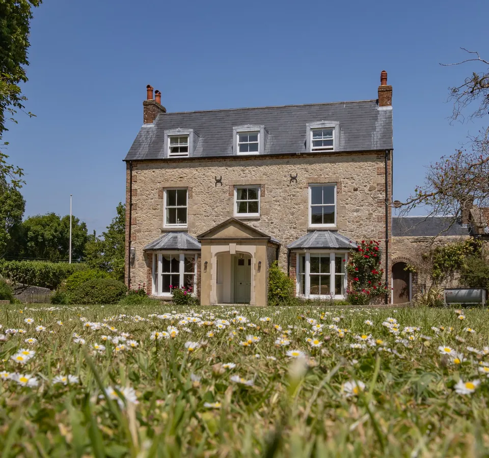 Stone farmhouse with slate roof and bay windows, viewed across a lawn dotted with daisies under a clear blue sky