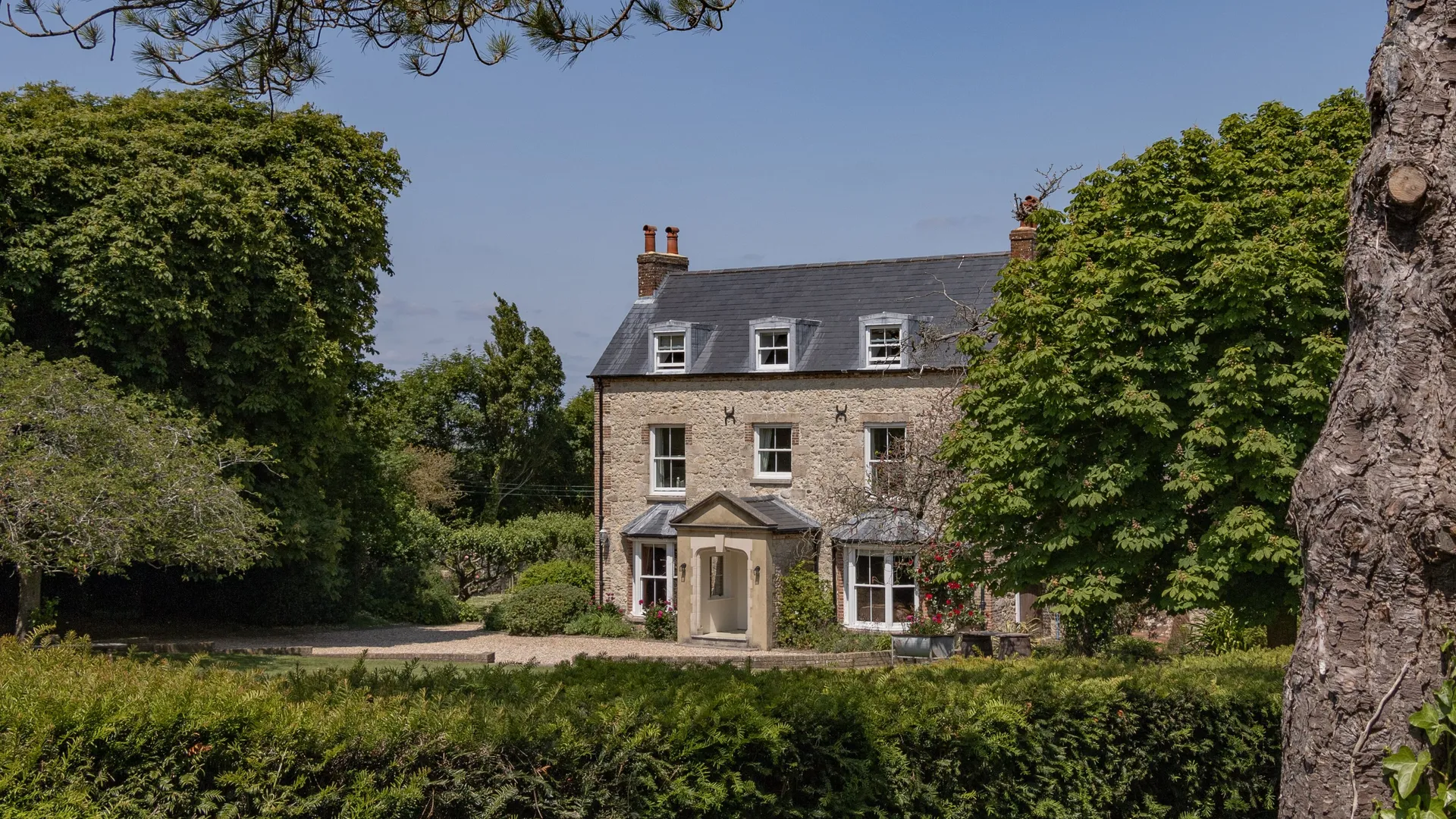 Stone farmhouse with slate roof and dormer windows, set among mature trees and garden greenery under a clear blue sky