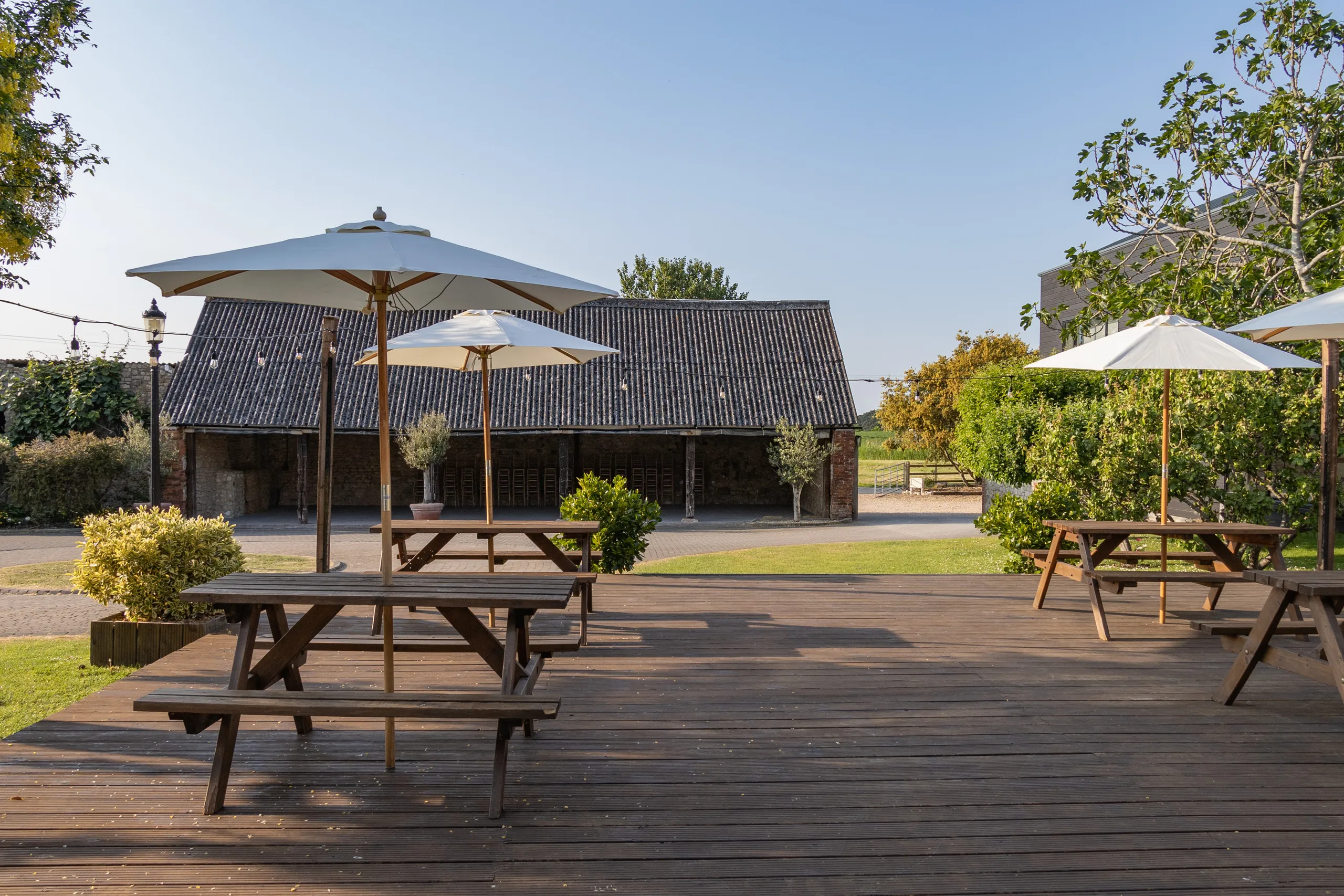 Outdoor wooden deck with picnic benches and white parasols facing a rustic barn