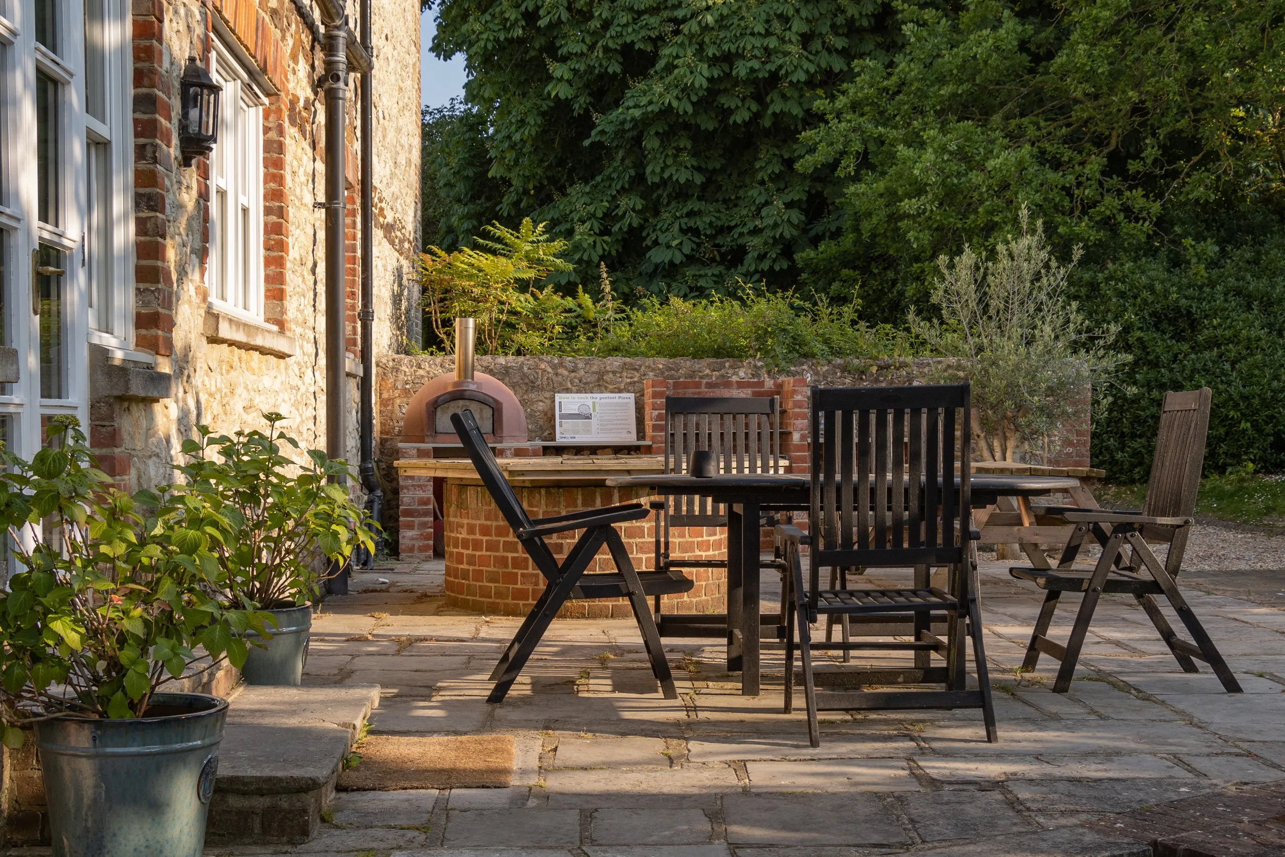 Patio seating and brick pizza oven beside a stone farmhouse in the evening sun