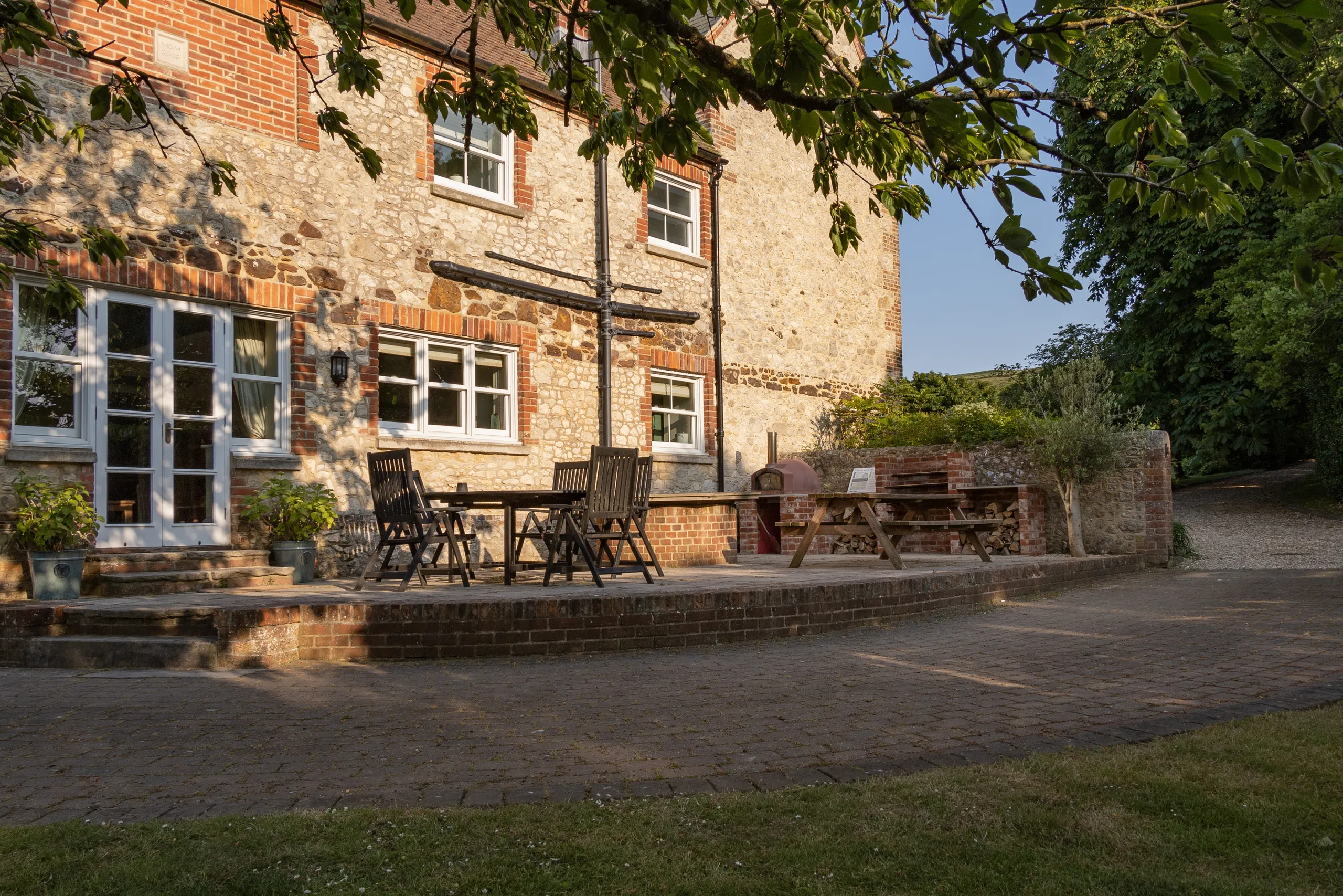 Stone and brick farmhouse patio with outdoor dining table, chairs and picnic bench in evening light