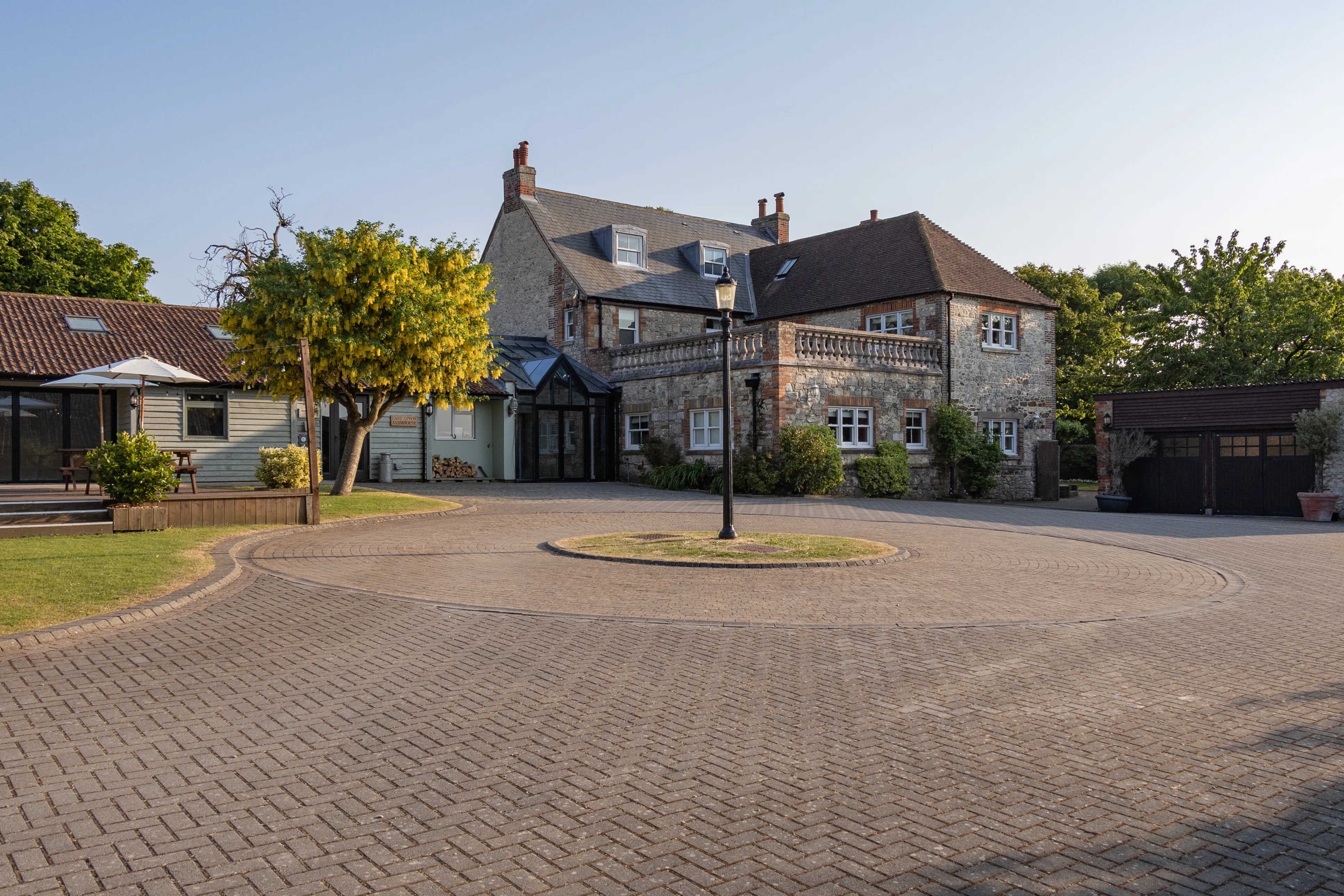 Stone farmhouse exterior with circular driveway, trees and outbuildings in warm evening light