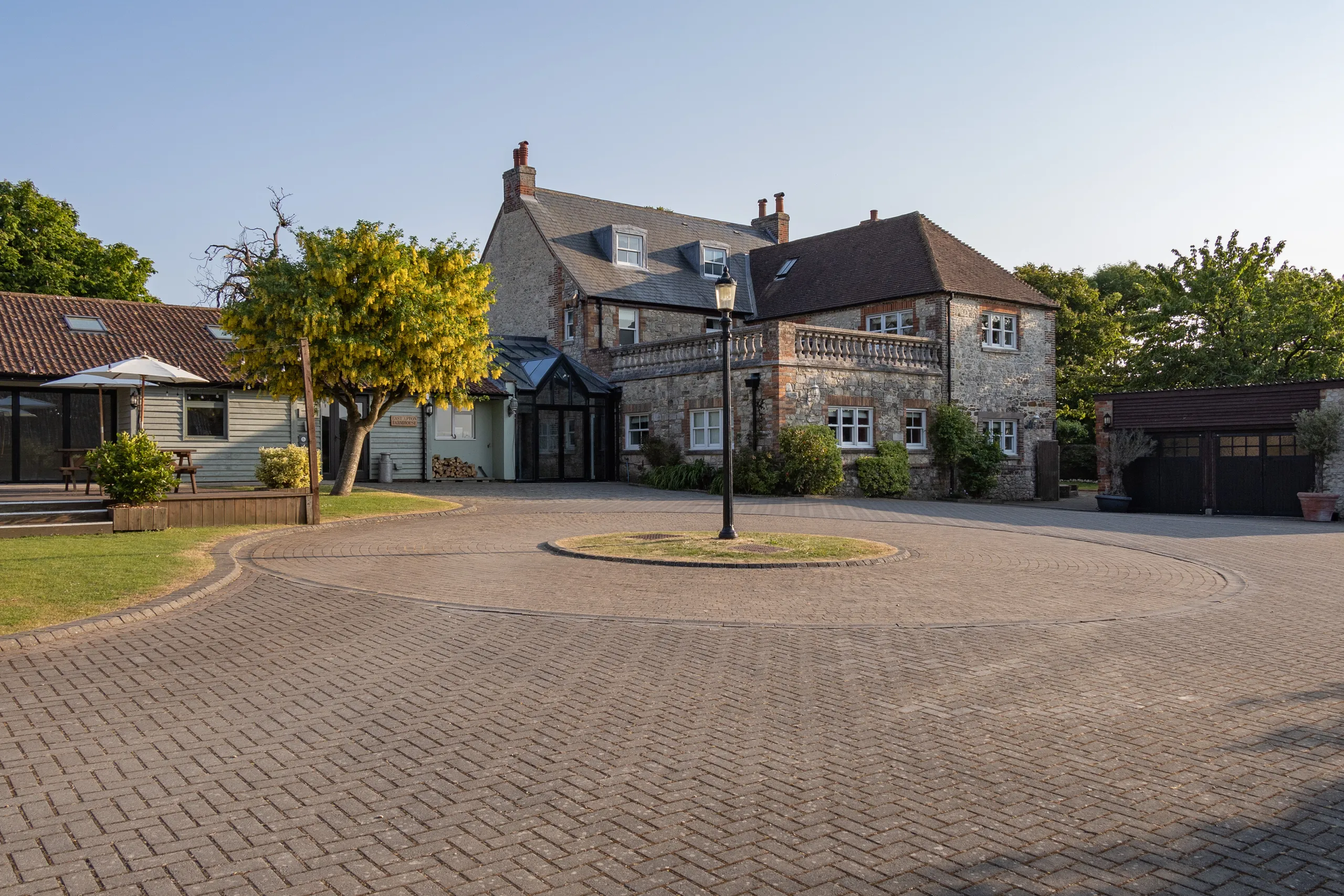 Stone farmhouse exterior with circular driveway, trees and outbuildings in warm evening light