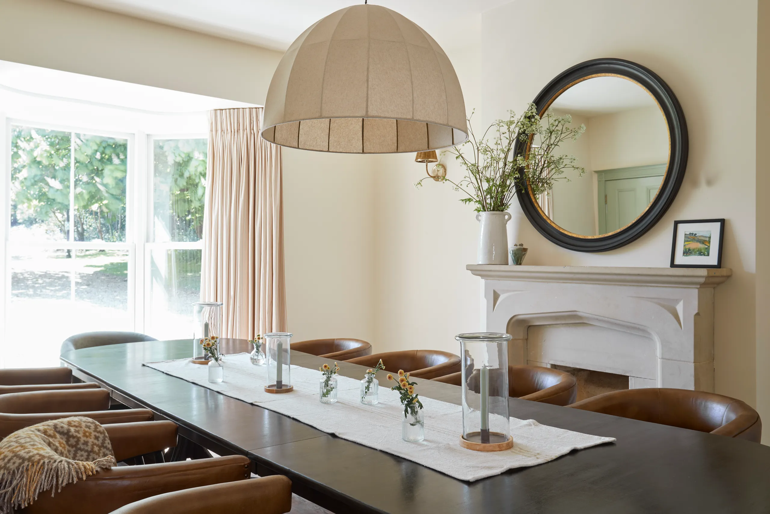 Bright farmhouse dining room with long table, tan leather chairs, large pendant light and round mirror above a stone fireplace