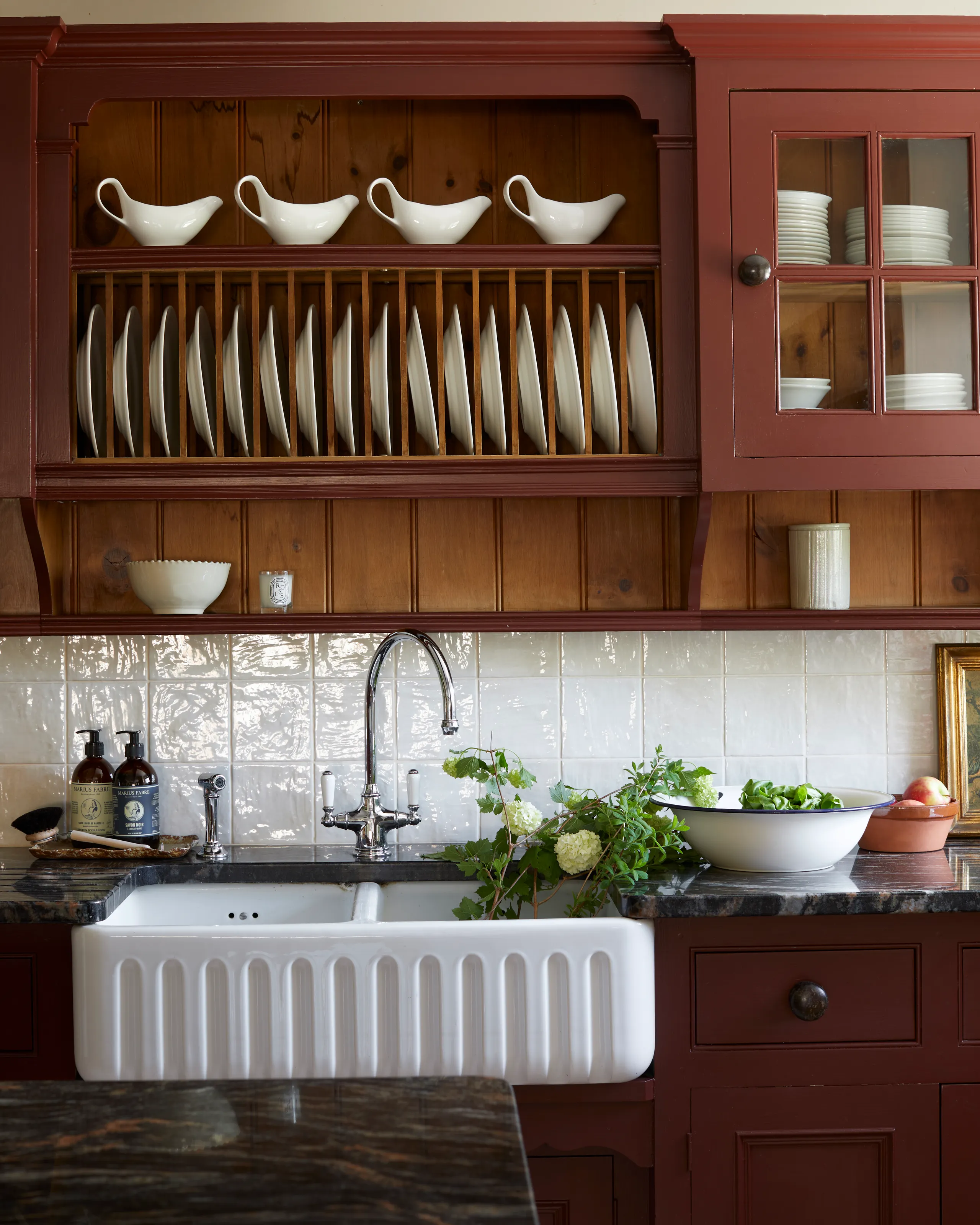 Farmhouse kitchen sink with deep white Belfast sink, dark worktop, white tiled splashback and burgundy plate rack filled with white crockery.