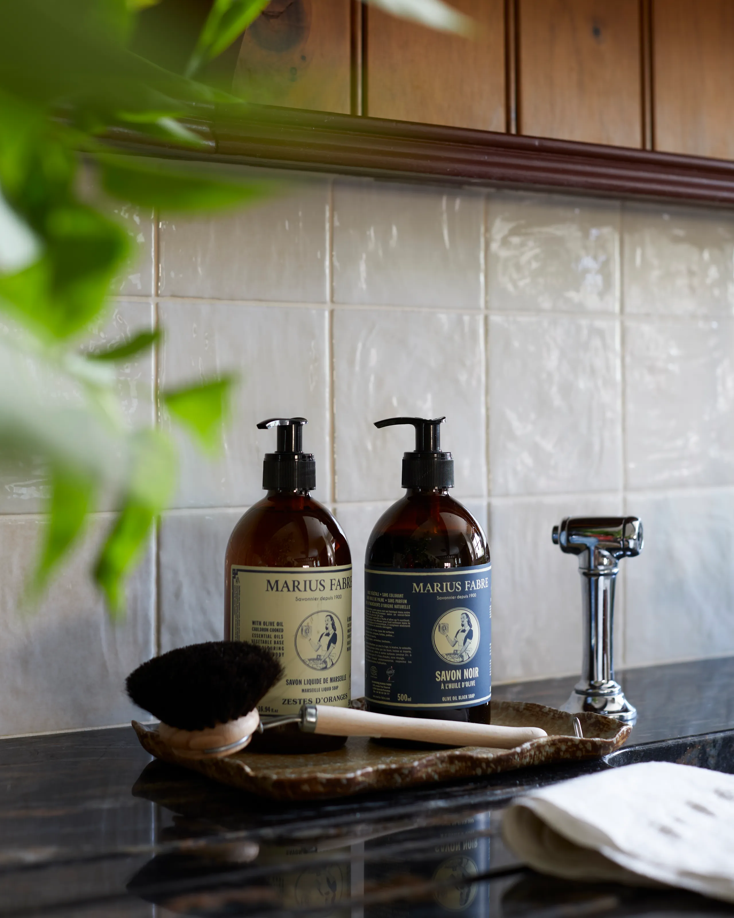 Two amber pump bottles of liquid soap beside a wooden dish brush on a tray, with a chrome tap and white tiled splashback.