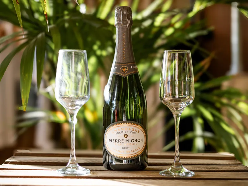Bottle of Pierre Mignon Champagne between two empty flutes on a wooden table with green foliage behind