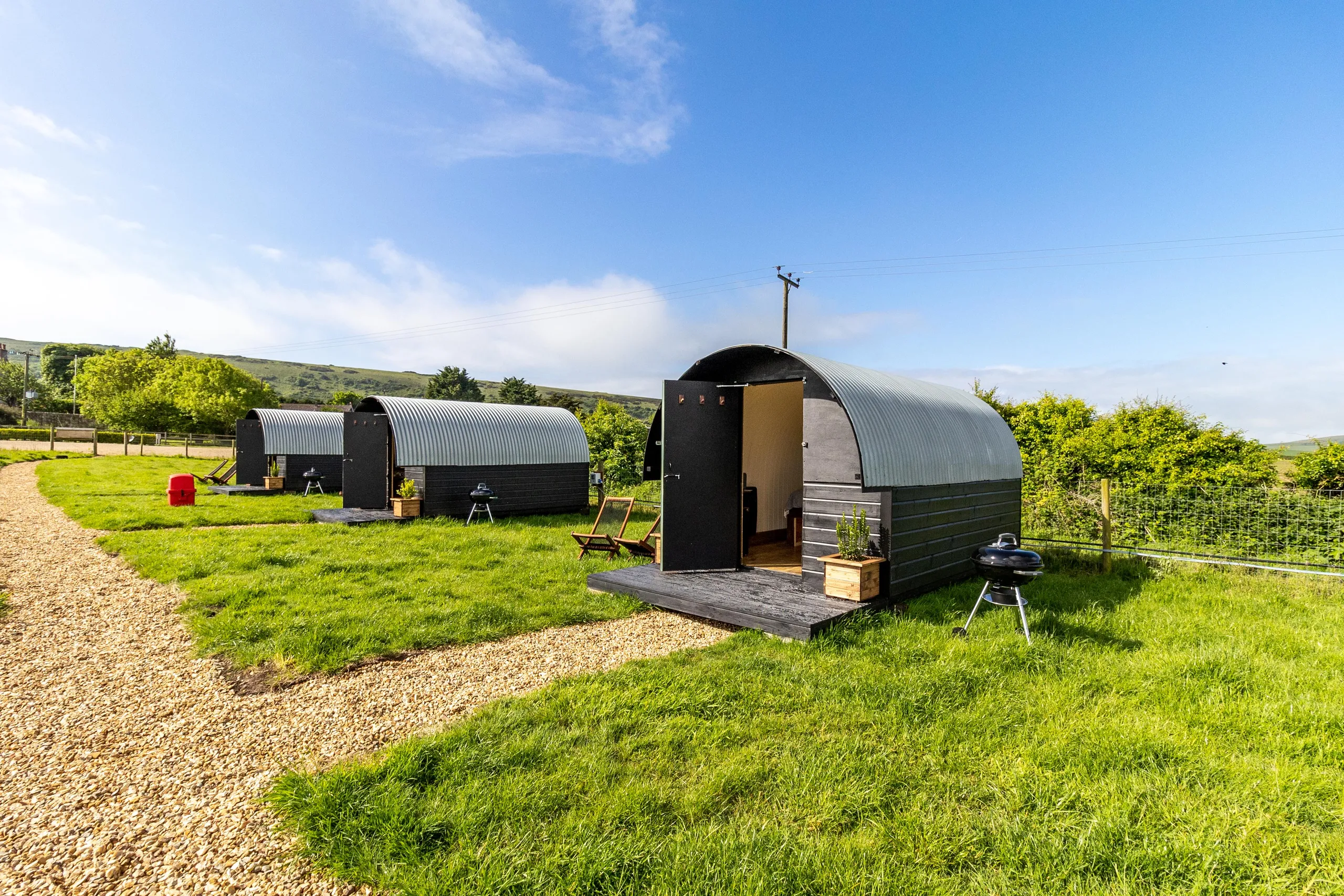 Row of modern shepherd’s hut pods with curved metal roofs on a grassy field under a blue sky