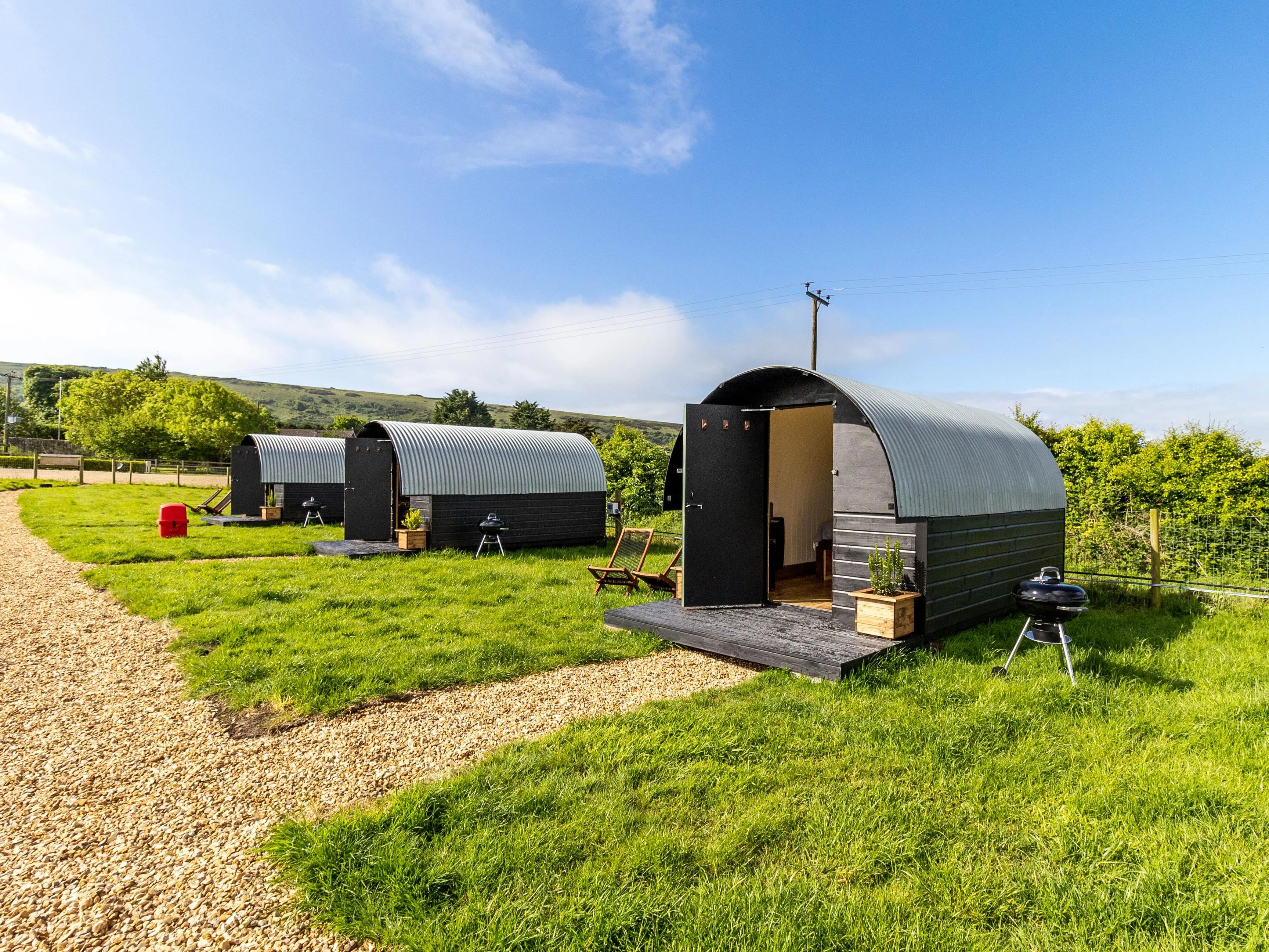 Row of modern shepherd’s hut pods with curved metal roofs on a grassy field under a blue sky