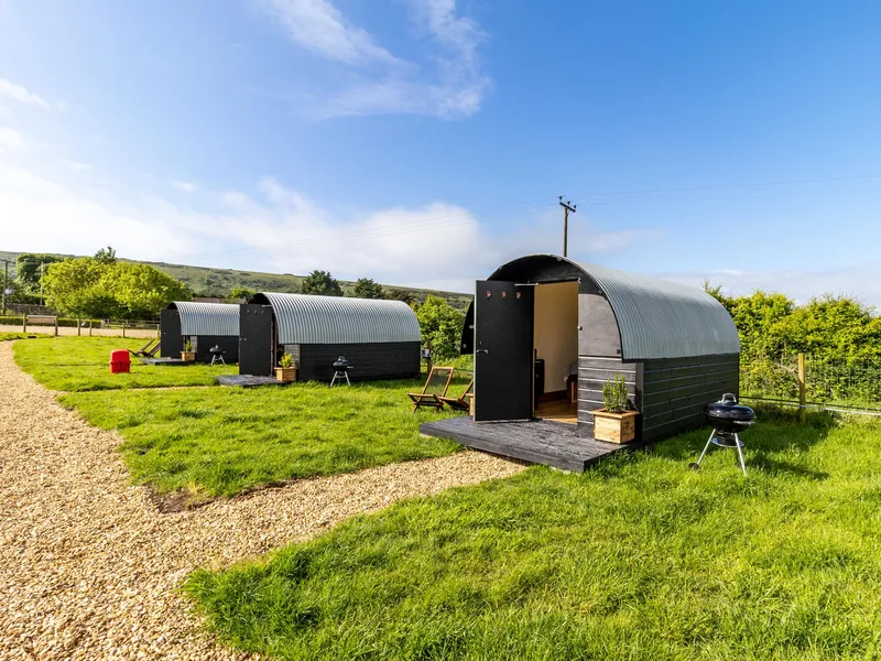 Row of modern shepherd’s hut pods with curved metal roofs on a grassy field under a blue sky