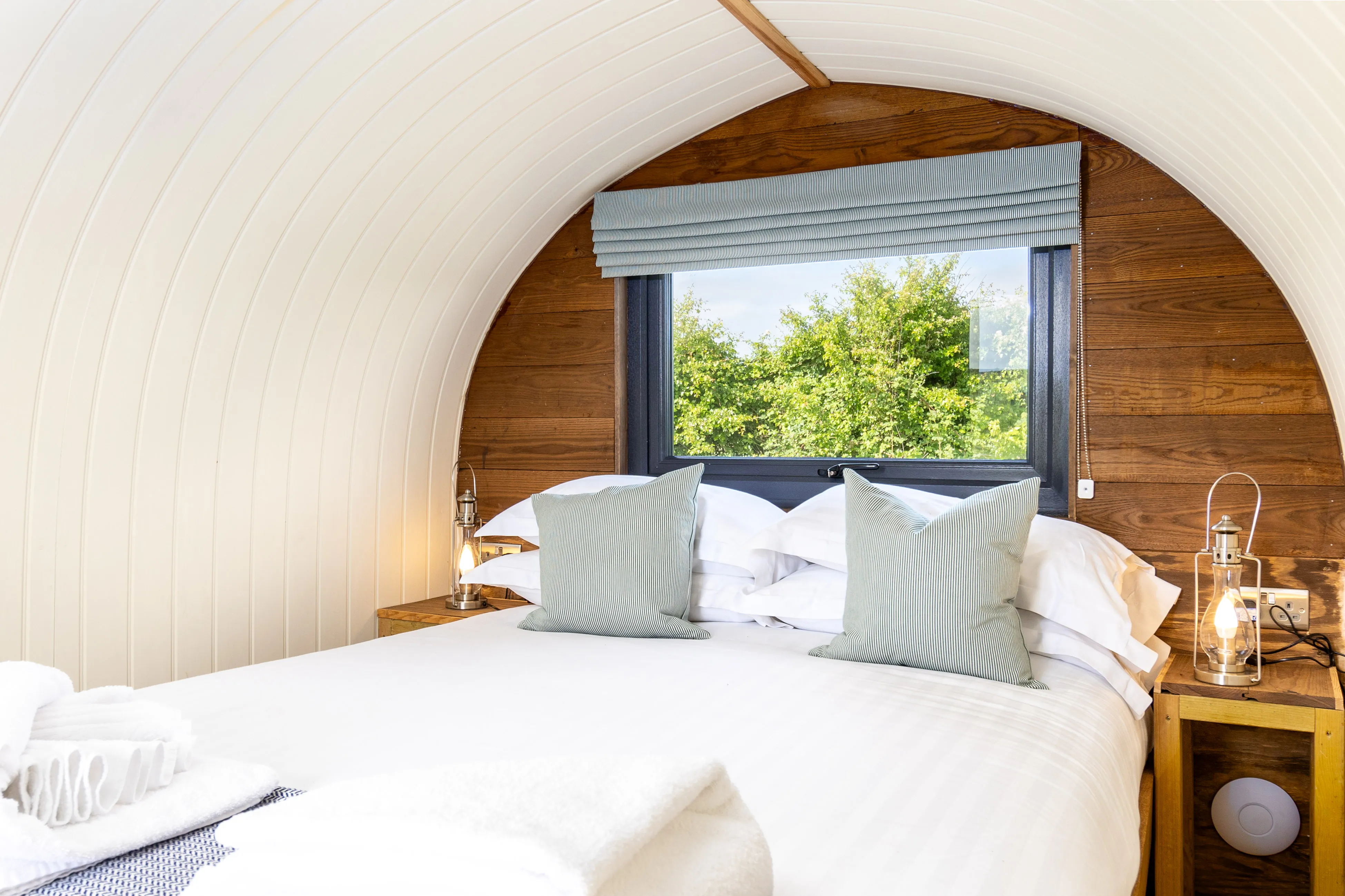 Cosy double bed inside a wooden shepherd’s hut with arched white ceiling, bedside lanterns and a window view of green trees