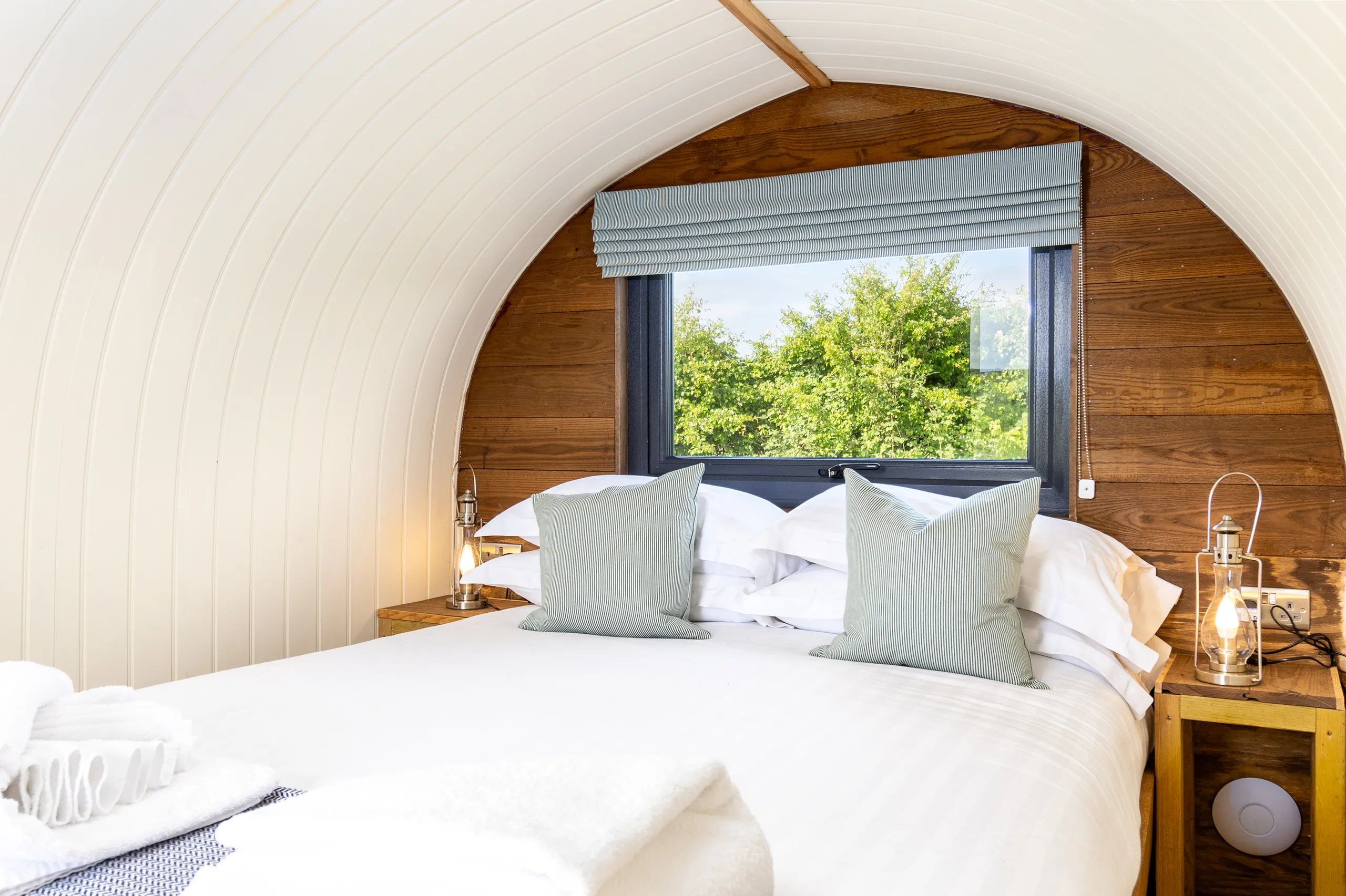 Cosy double bed inside a wooden shepherd’s hut with arched white ceiling, bedside lanterns and a window view of green trees