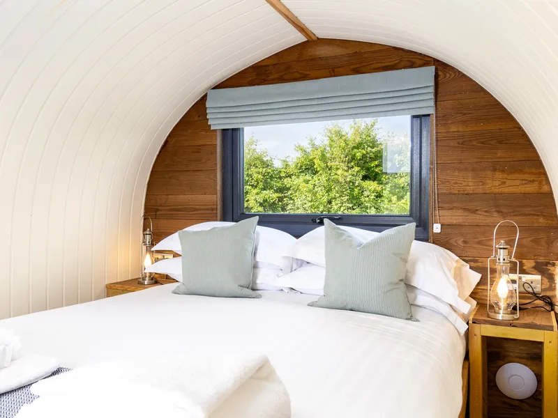Cosy double bed inside a wooden shepherd’s hut with arched white ceiling, bedside lanterns and a window view of green trees