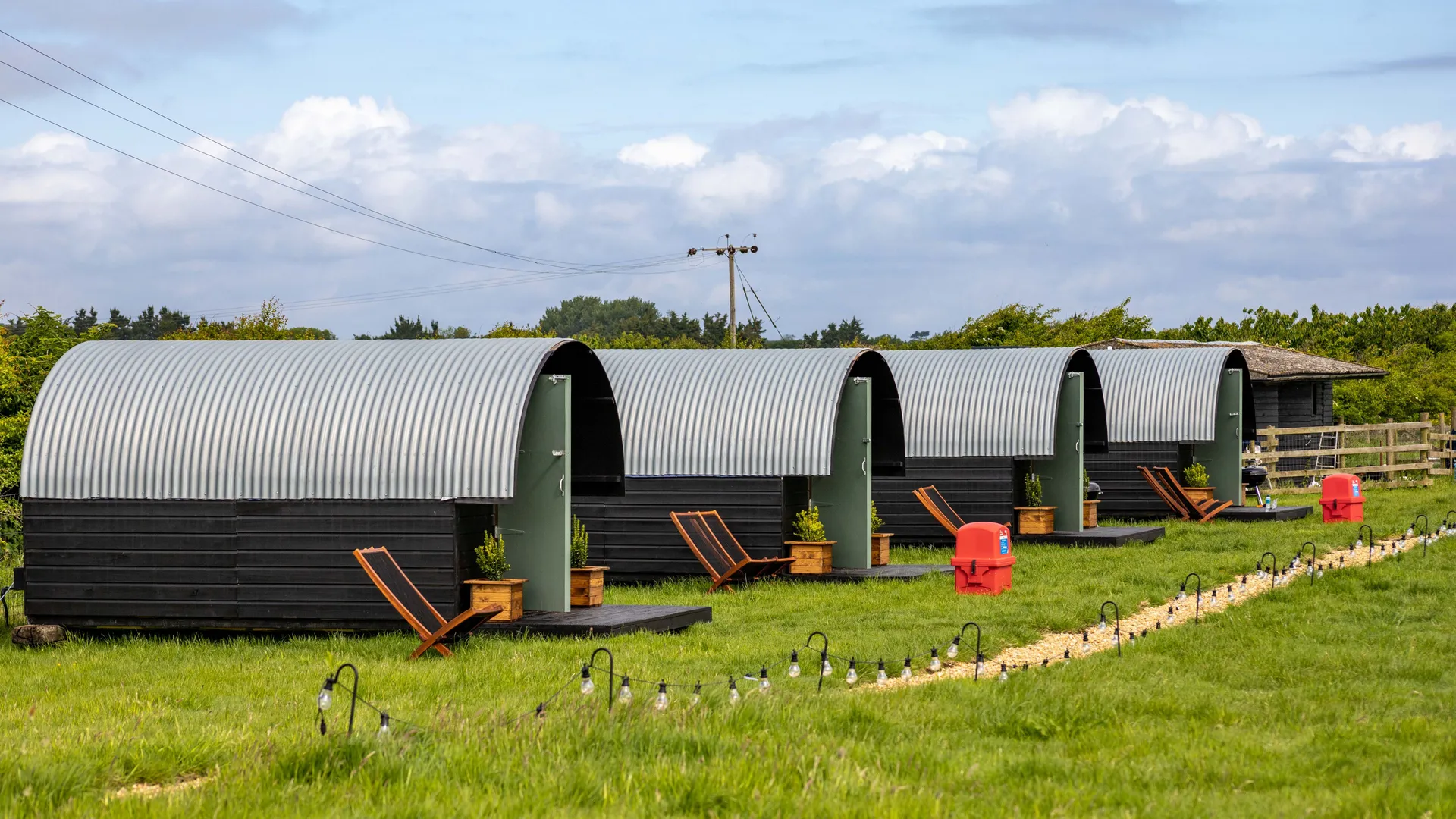 Row of corrugated metal shepherd’s huts on a grassy campsite with deckchairs and string lights