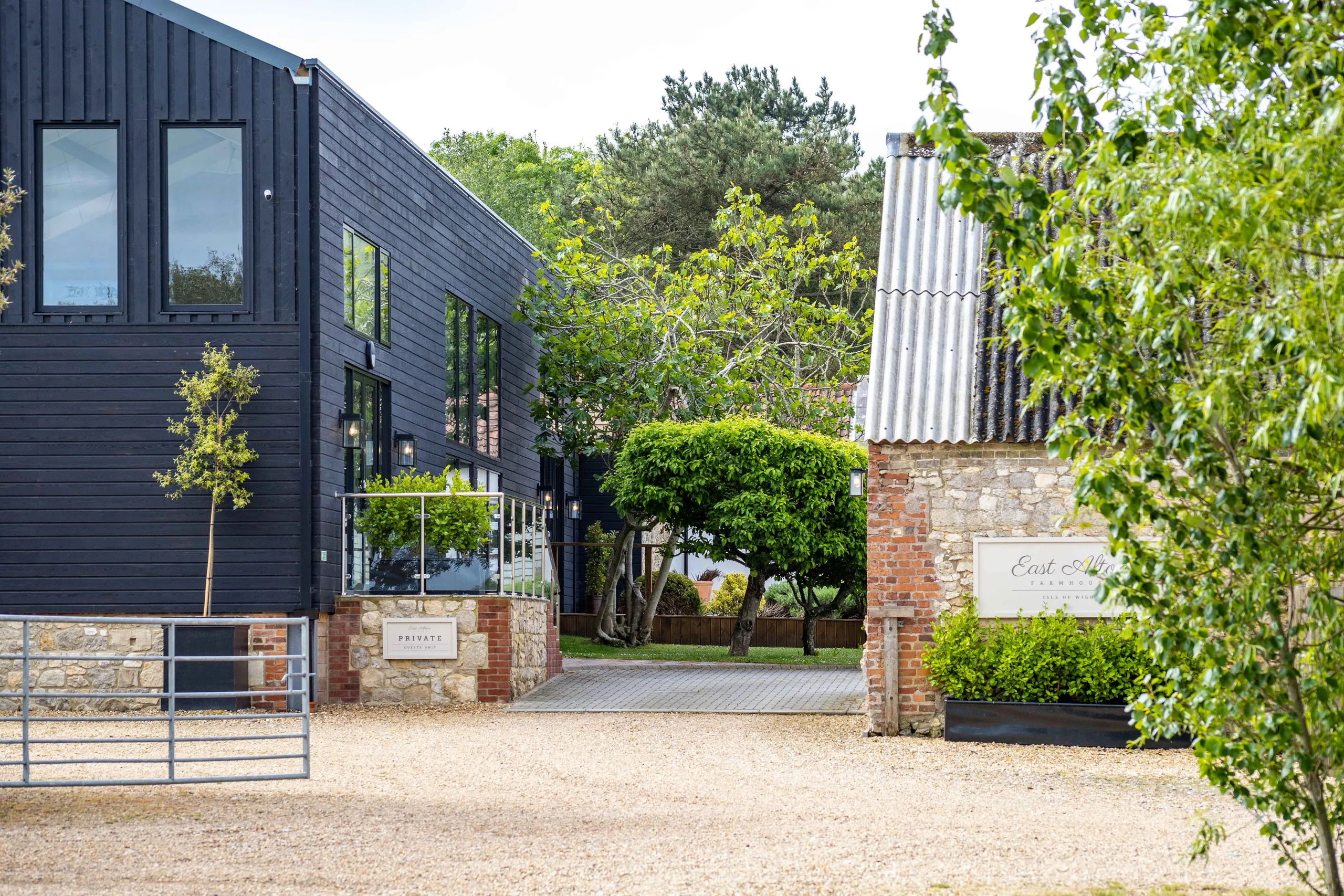 Modern black-clad building beside a brick-and-stone entrance with trees and a gravel driveway