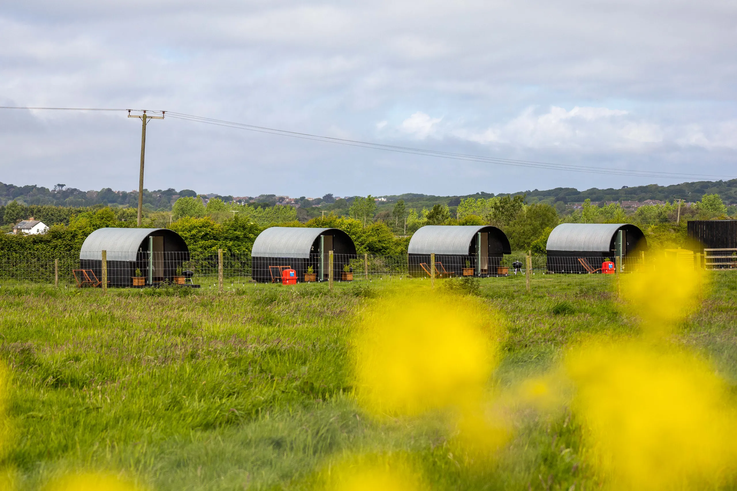 Four arched glamping huts in a grassy field with wildflowers under a cloudy sky