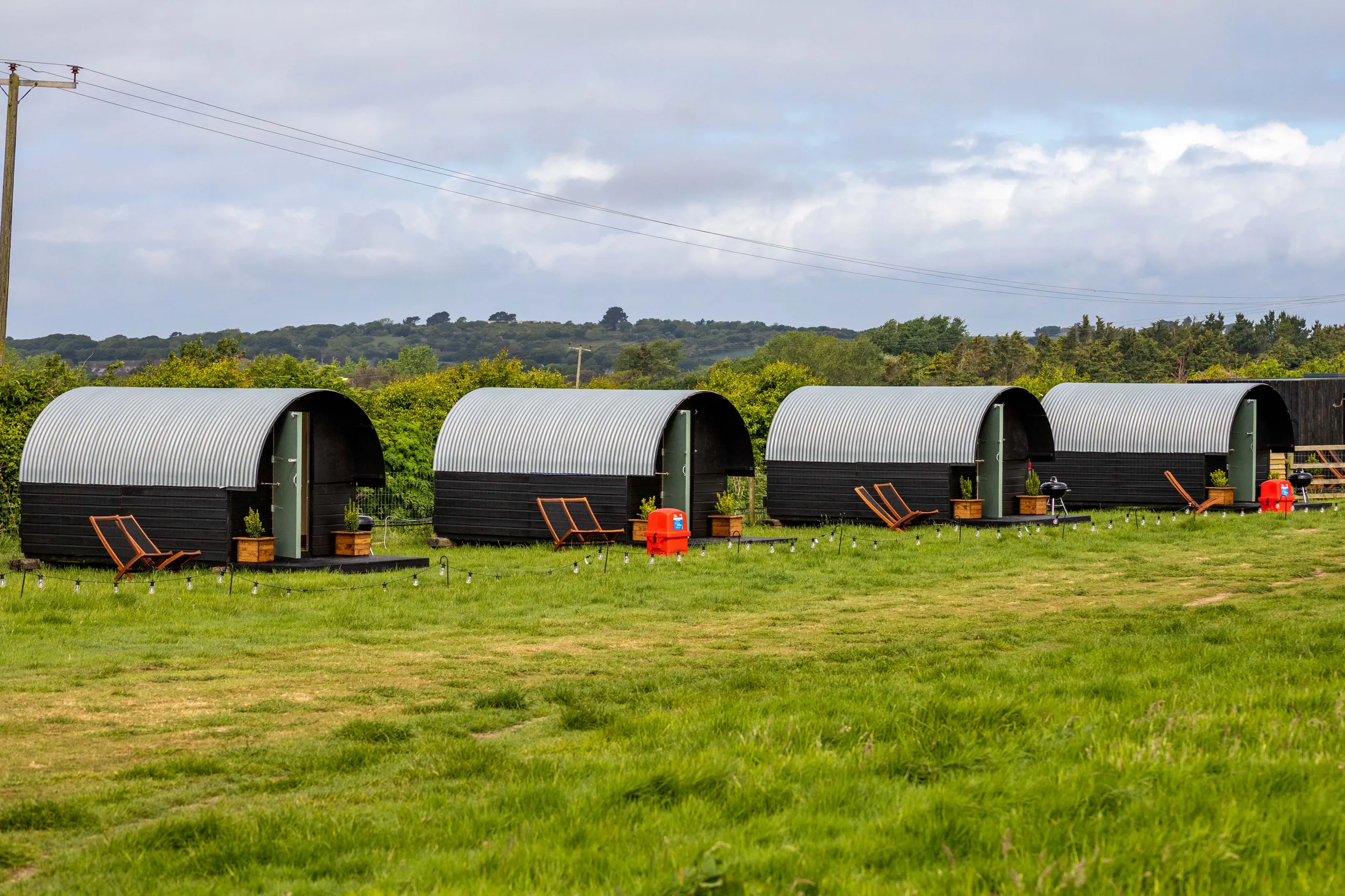 Row of four curved, corrugated hut cabins on a grassy field with deckchairs and outdoor lights