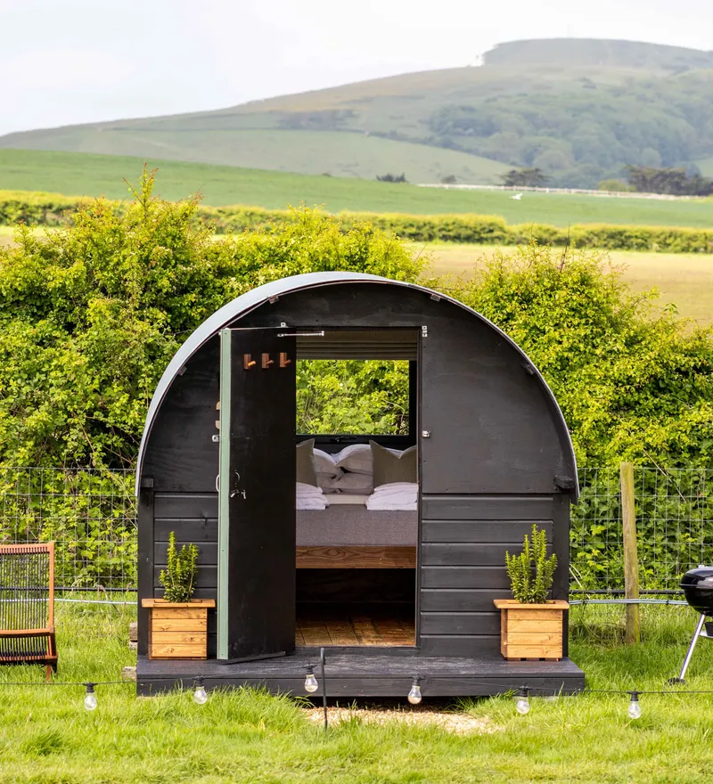 Cosy black shepherd’s hut with open door, deckchairs and a barbecue in a green countryside field