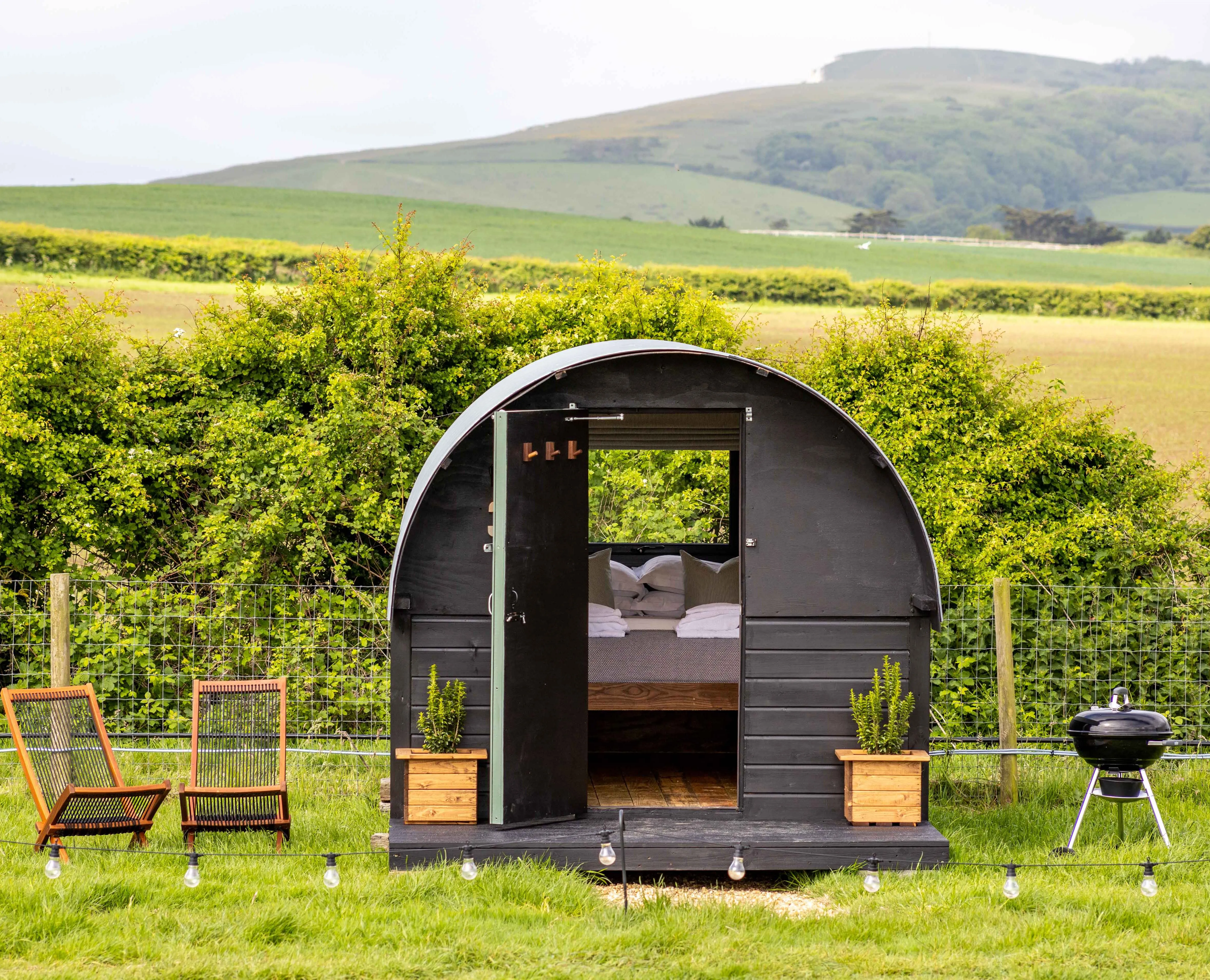 Cosy black shepherd’s hut with open door, deckchairs and a barbecue in a green countryside field
