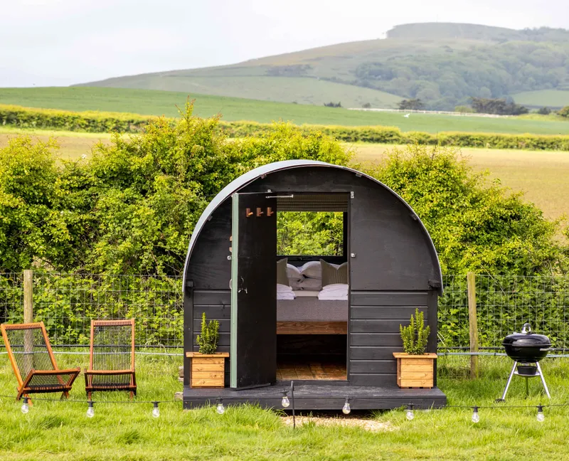 Cosy black shepherd’s hut with open door, deckchairs and a barbecue in a green countryside field