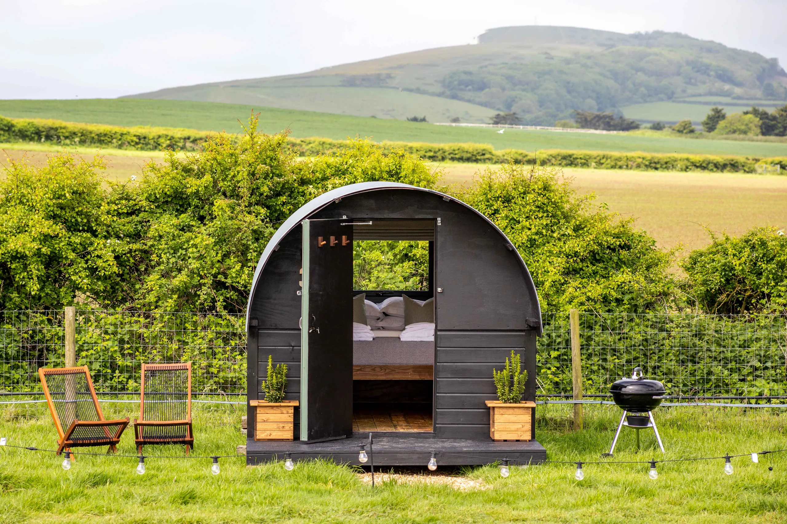 Cosy black shepherd’s hut with open door, deckchairs and a barbecue in a green countryside field