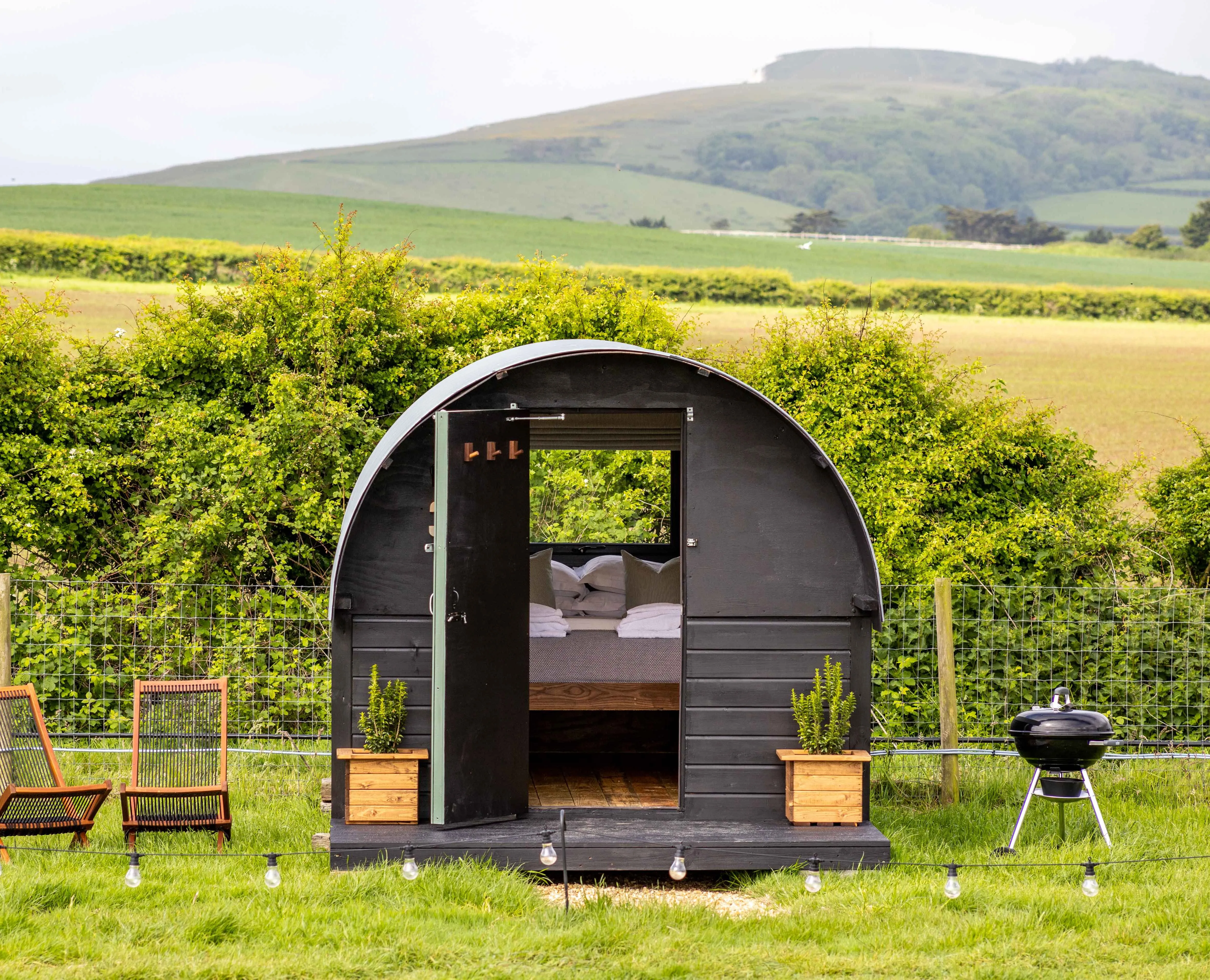Cosy black shepherd’s hut with open door, deckchairs and a barbecue in a green countryside field