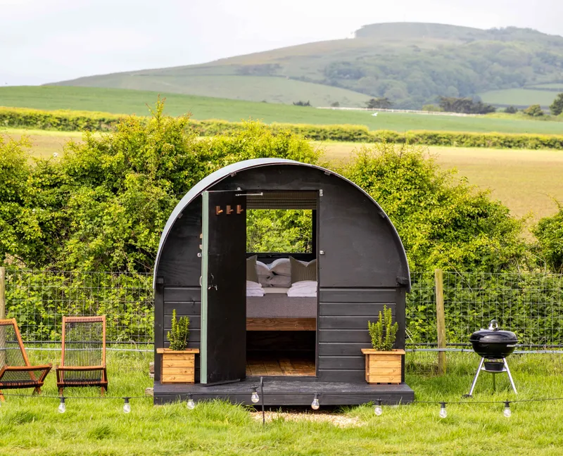 Cosy black shepherd’s hut with open door, deckchairs and a barbecue in a green countryside field