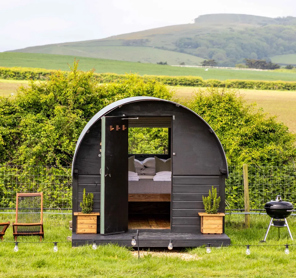 Cosy black shepherd’s hut with open door, deckchairs and a barbecue in a green countryside field