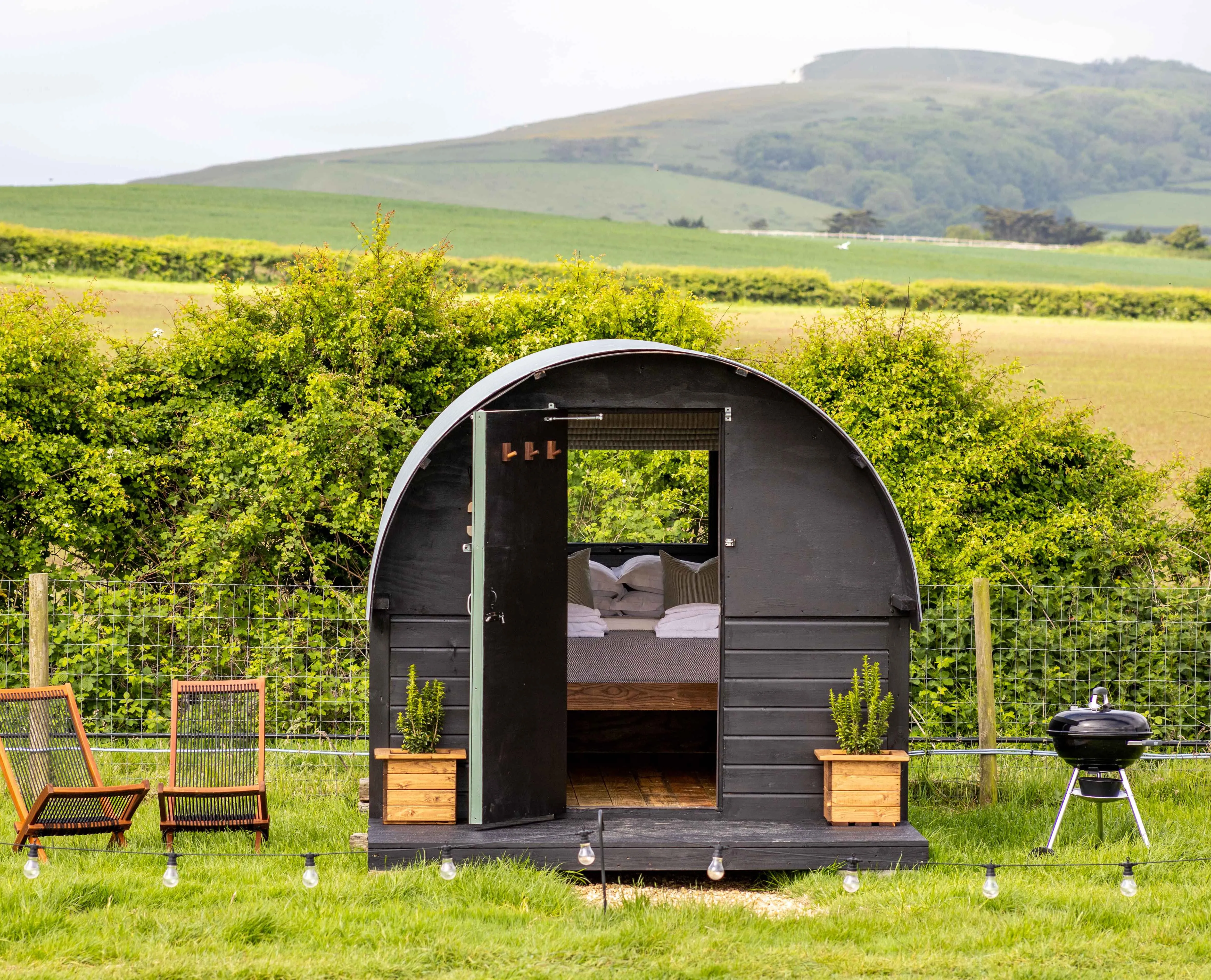 Cosy black shepherd’s hut with open door, deckchairs and a barbecue in a green countryside field