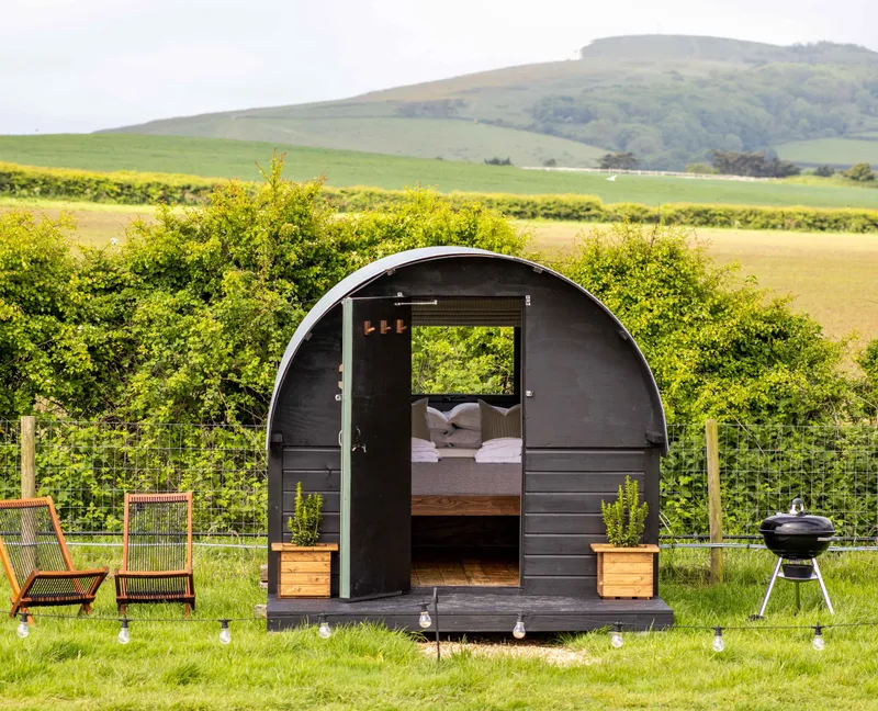 Cosy black shepherd’s hut with open door, deckchairs and a barbecue in a green countryside field